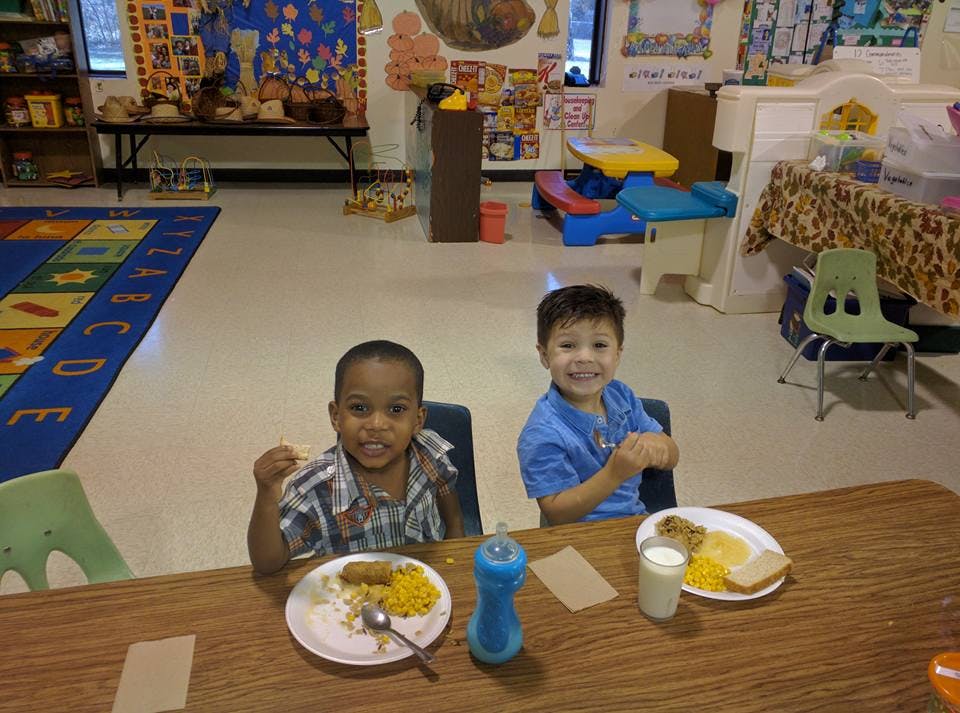 Two children sitting at a table with plates of food in a classroom