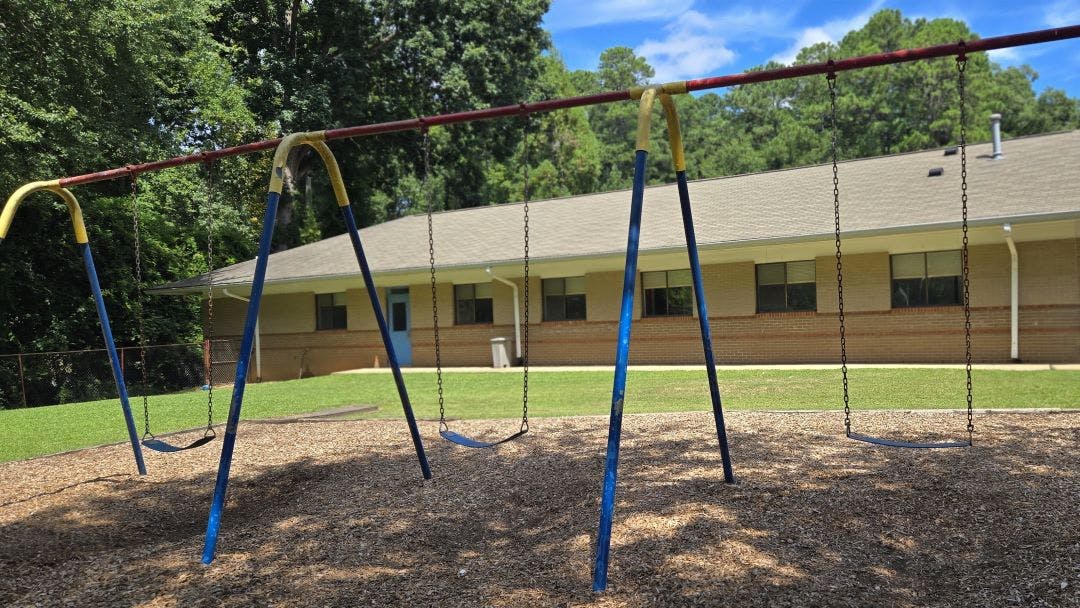 Empty swing set on wood chips in front of a brick building and trees