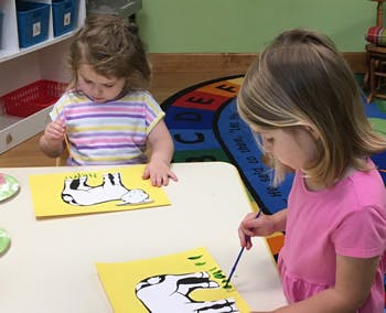 Two children painting cow cutouts on yellow paper at a table