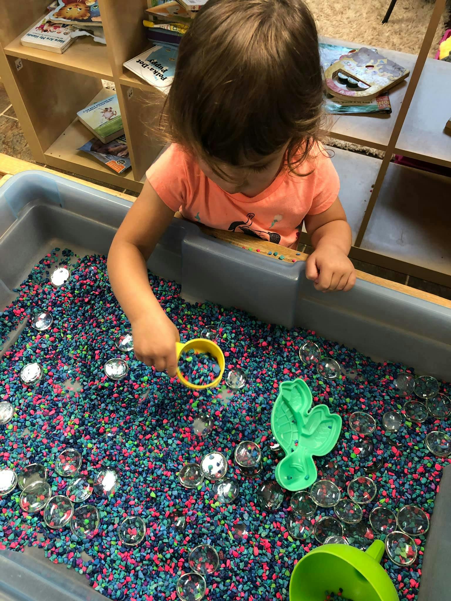 Child playing with a yellow scoop in a sensory bin filled with colorful pebbles