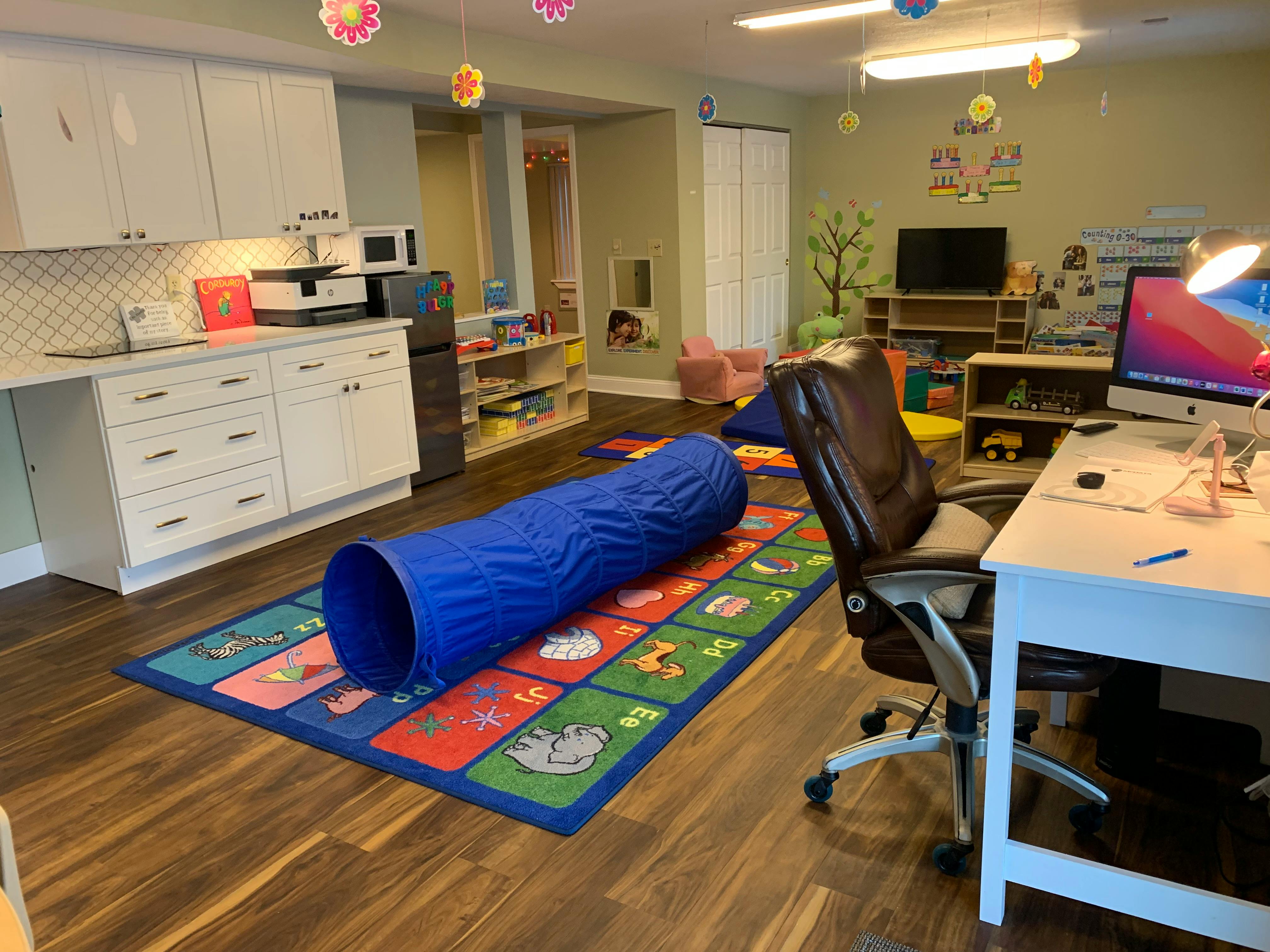 Playroom with a blue tunnel on a colorful rug, desk with computer, and kitchen cabinets