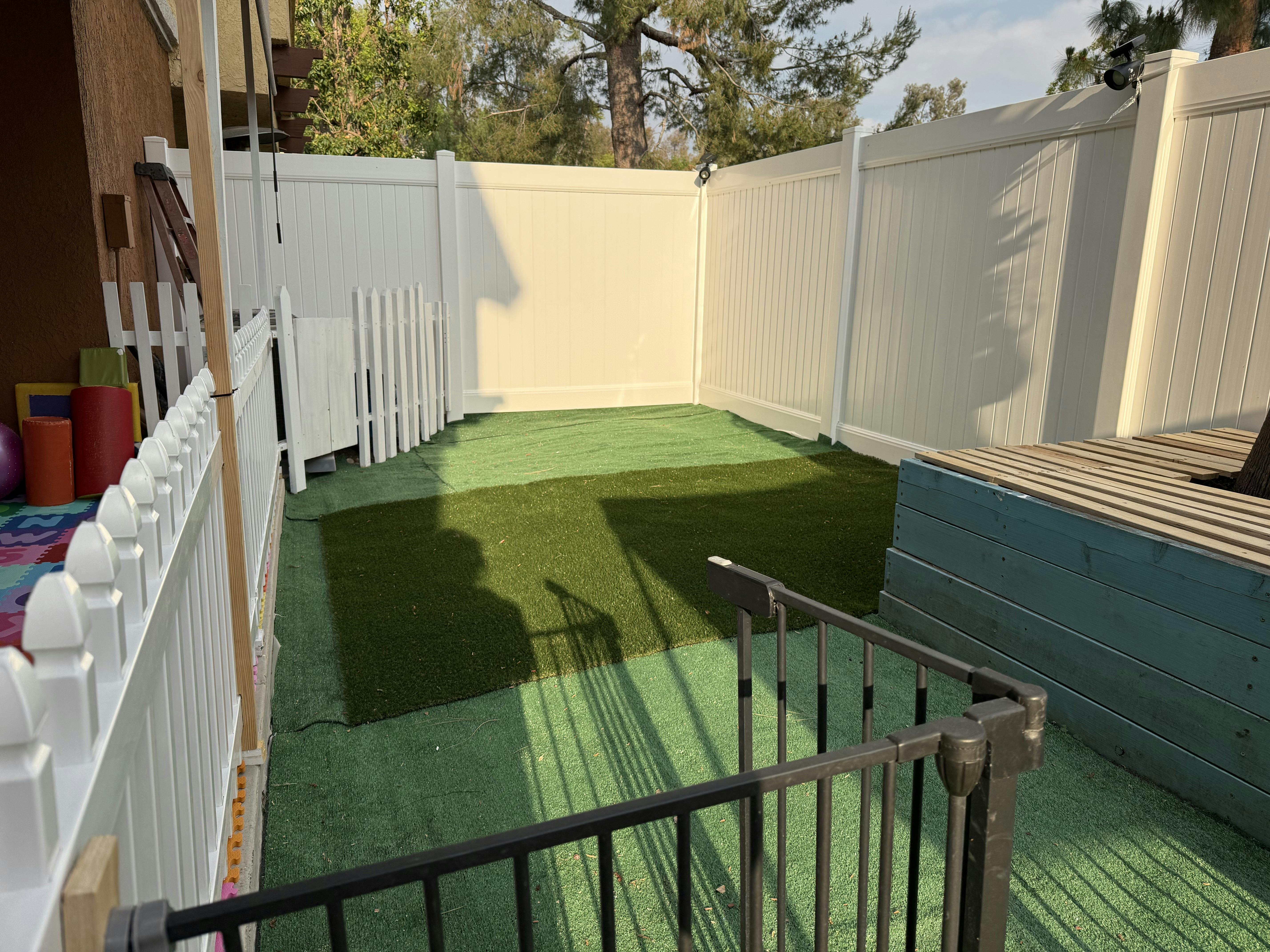 backyard patio with artificial turf, white picket fence, and dark railing