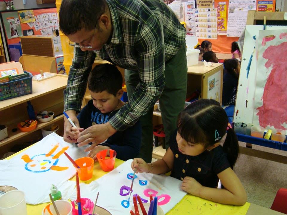 Adult helping a child paint at a table with another child