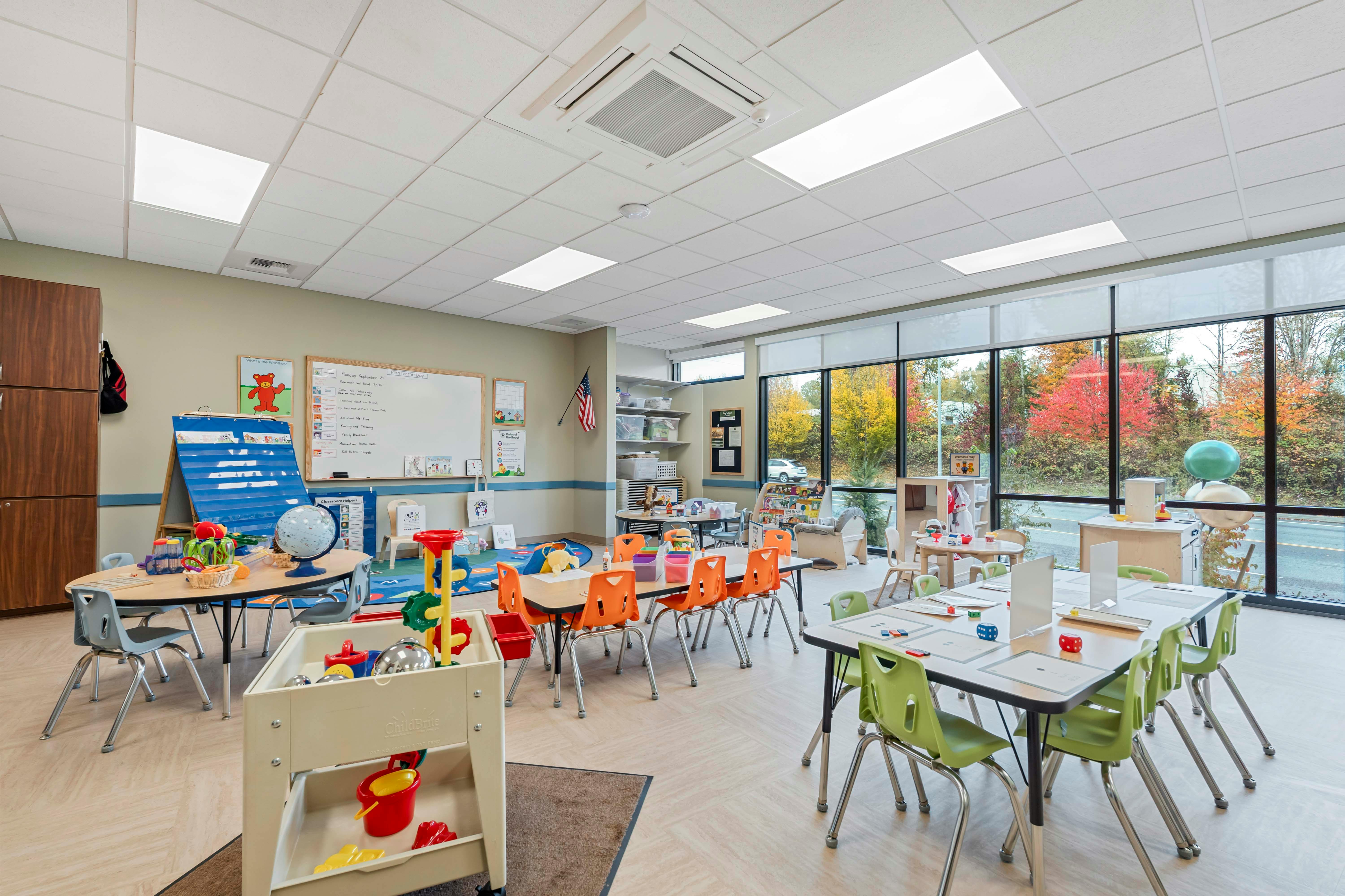 Colorful classroom with several small tables and chairs, toys, and a large window