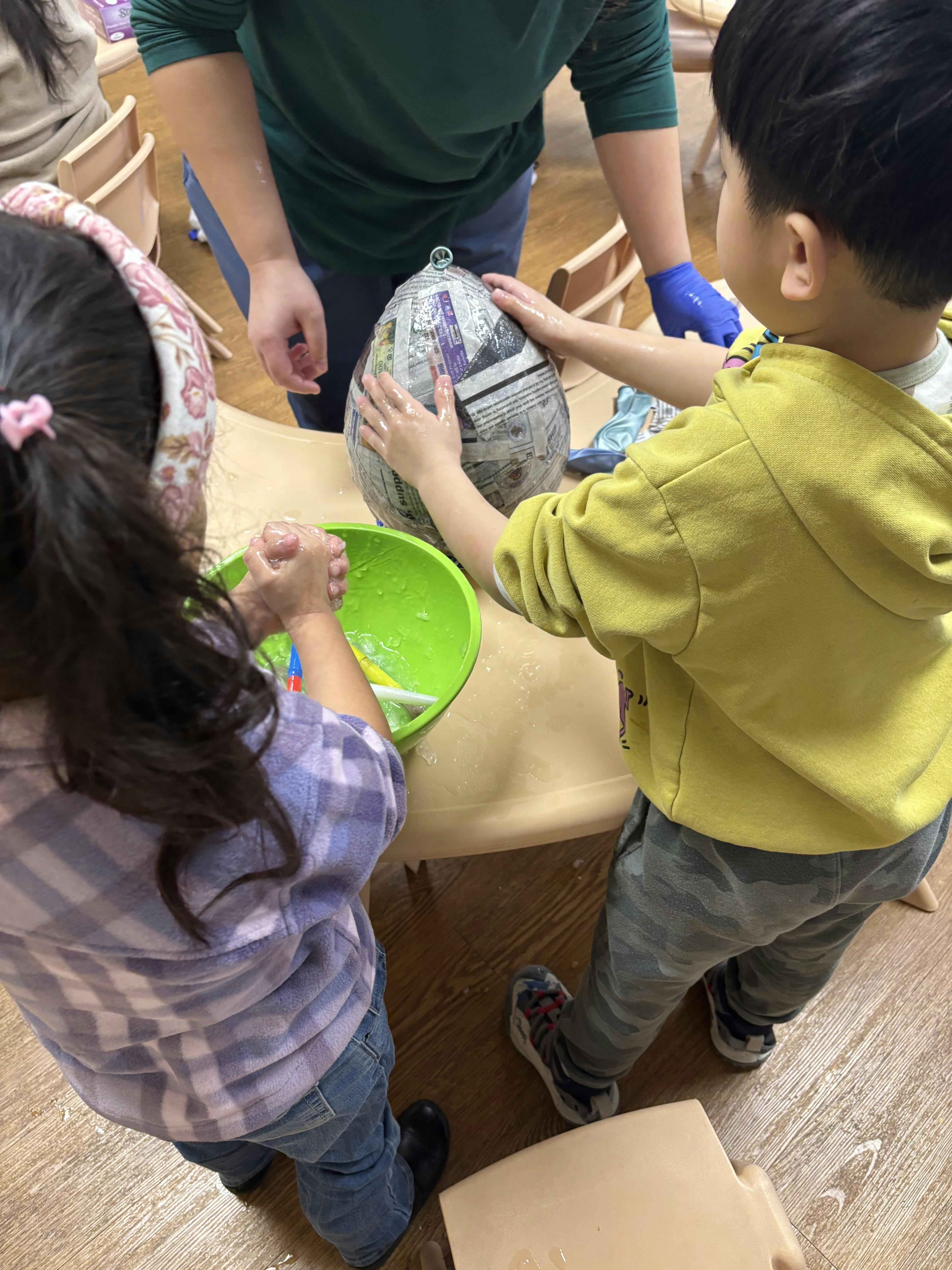 A person helping two children paper mache a balloon on a table