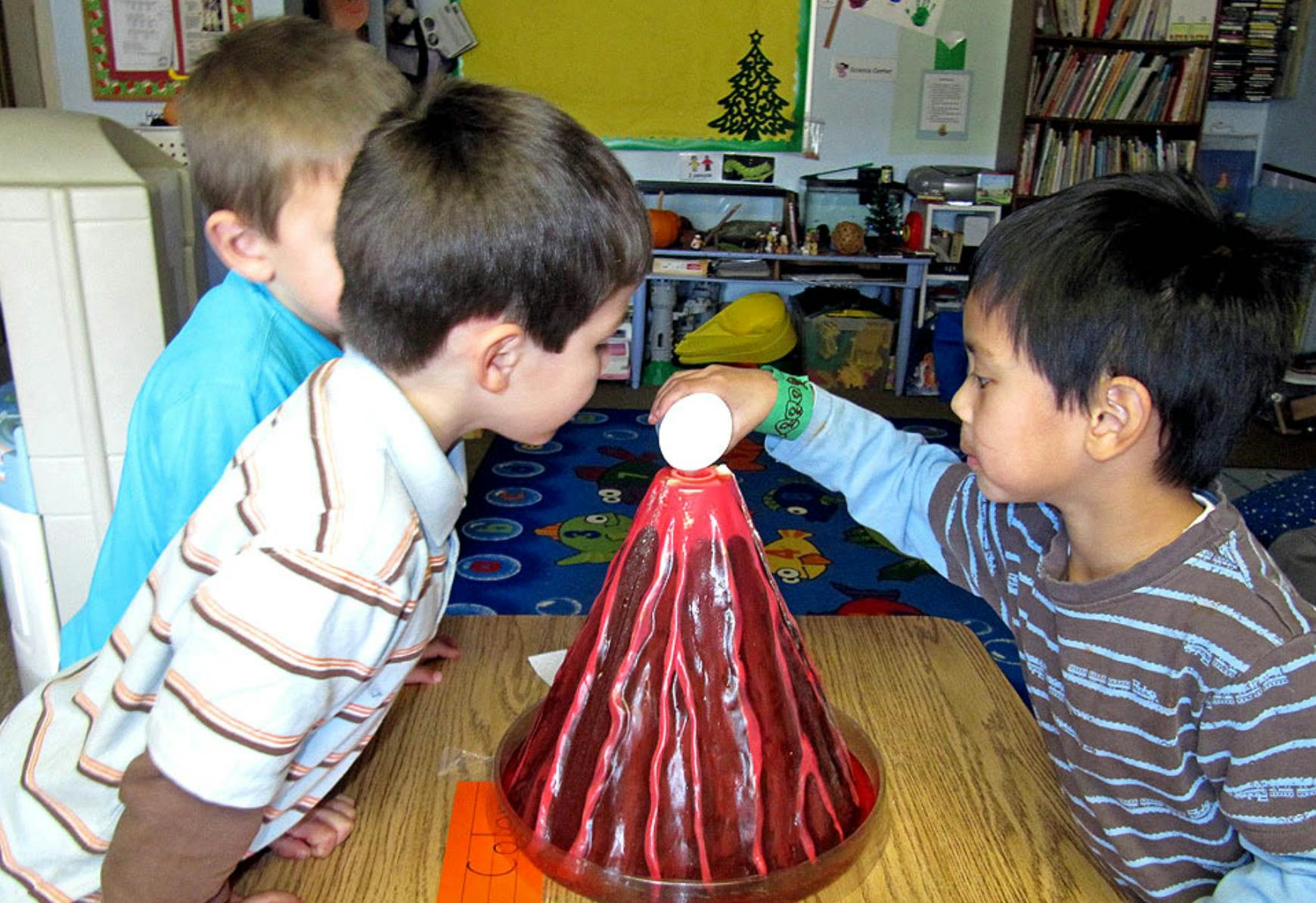 Three children observing a volcano model, one placing an object into it