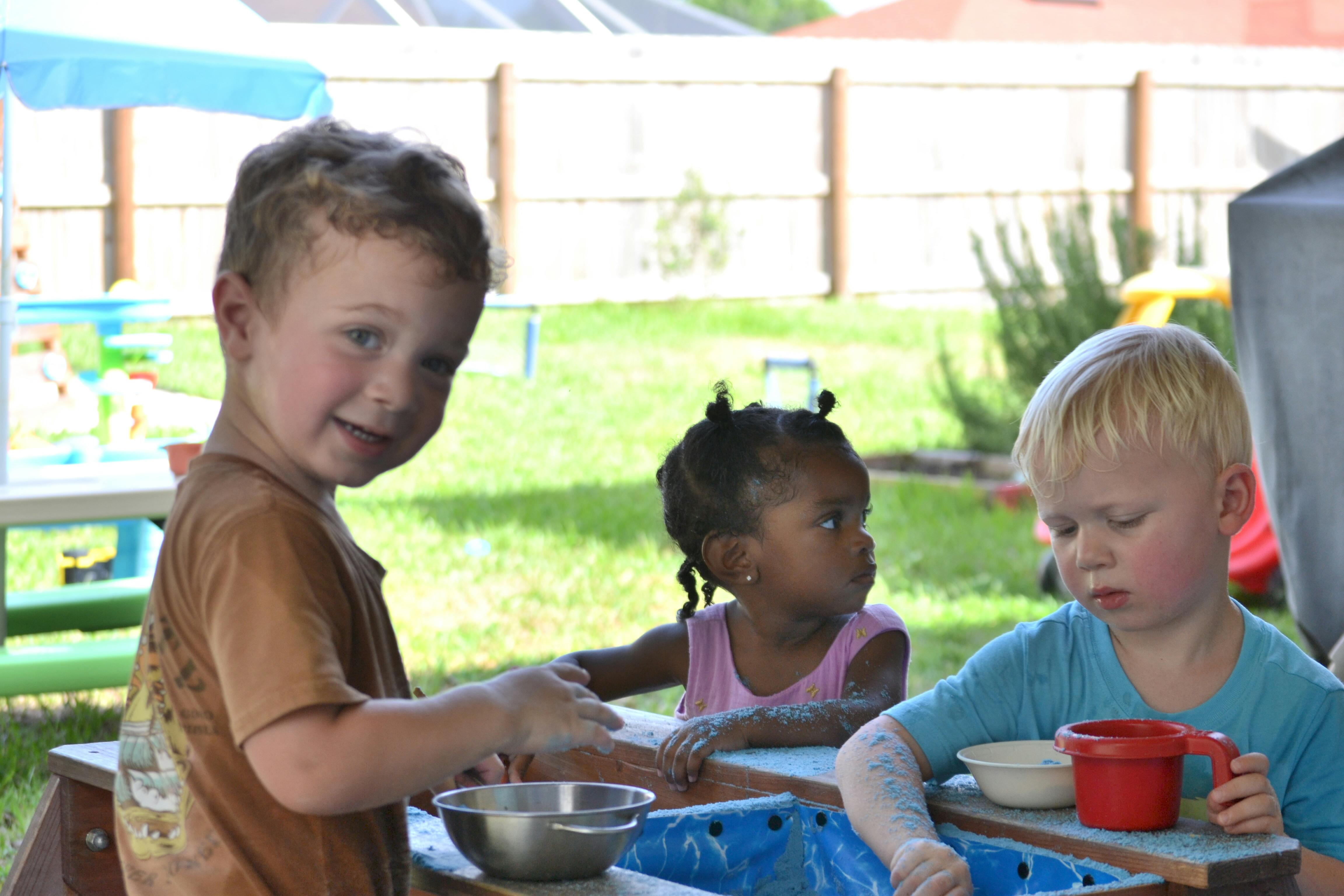 Three children playing with bowls and cups at a table outdoors
