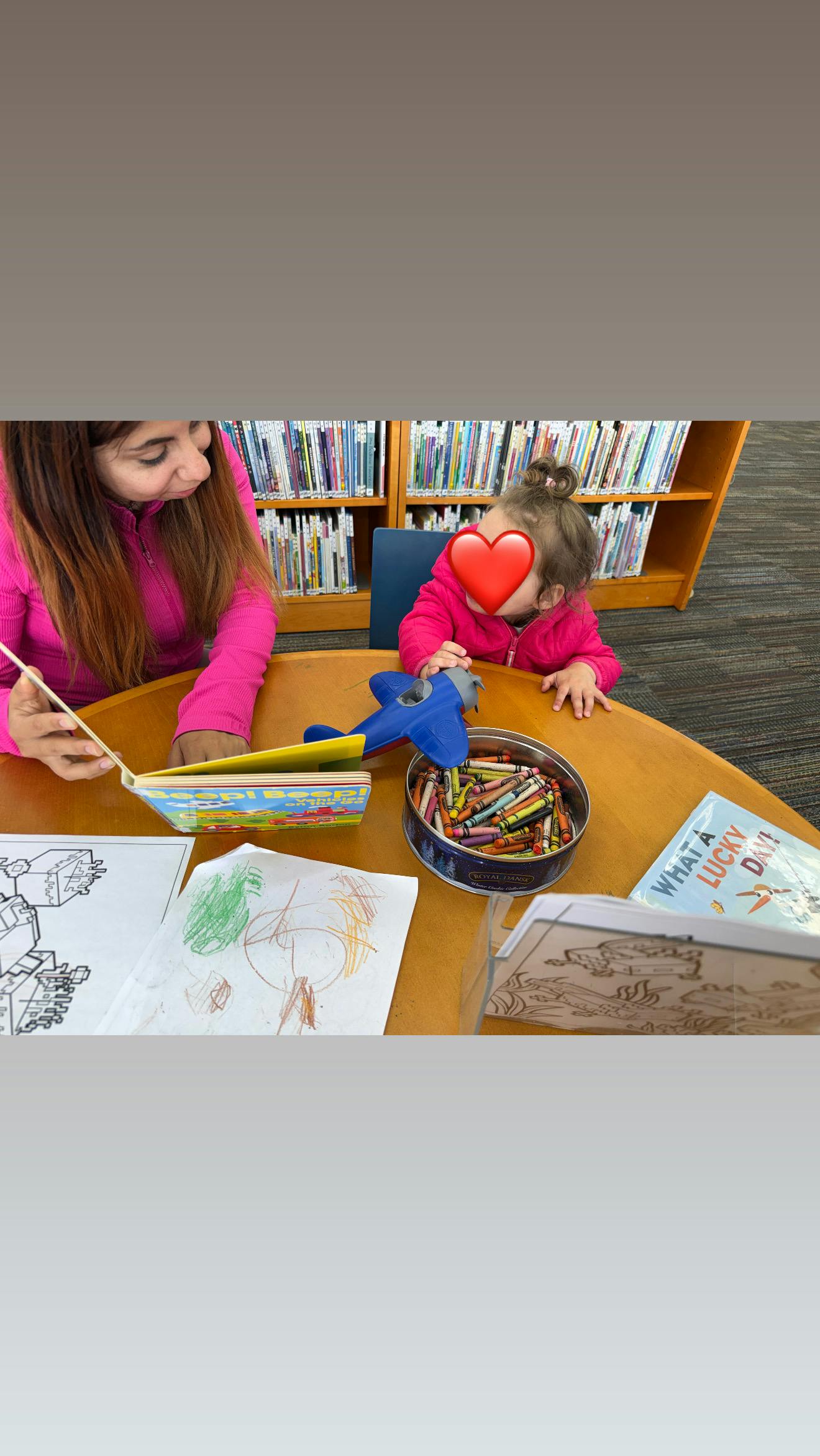 A person reading a book to a child at a library table with crayons and a toy