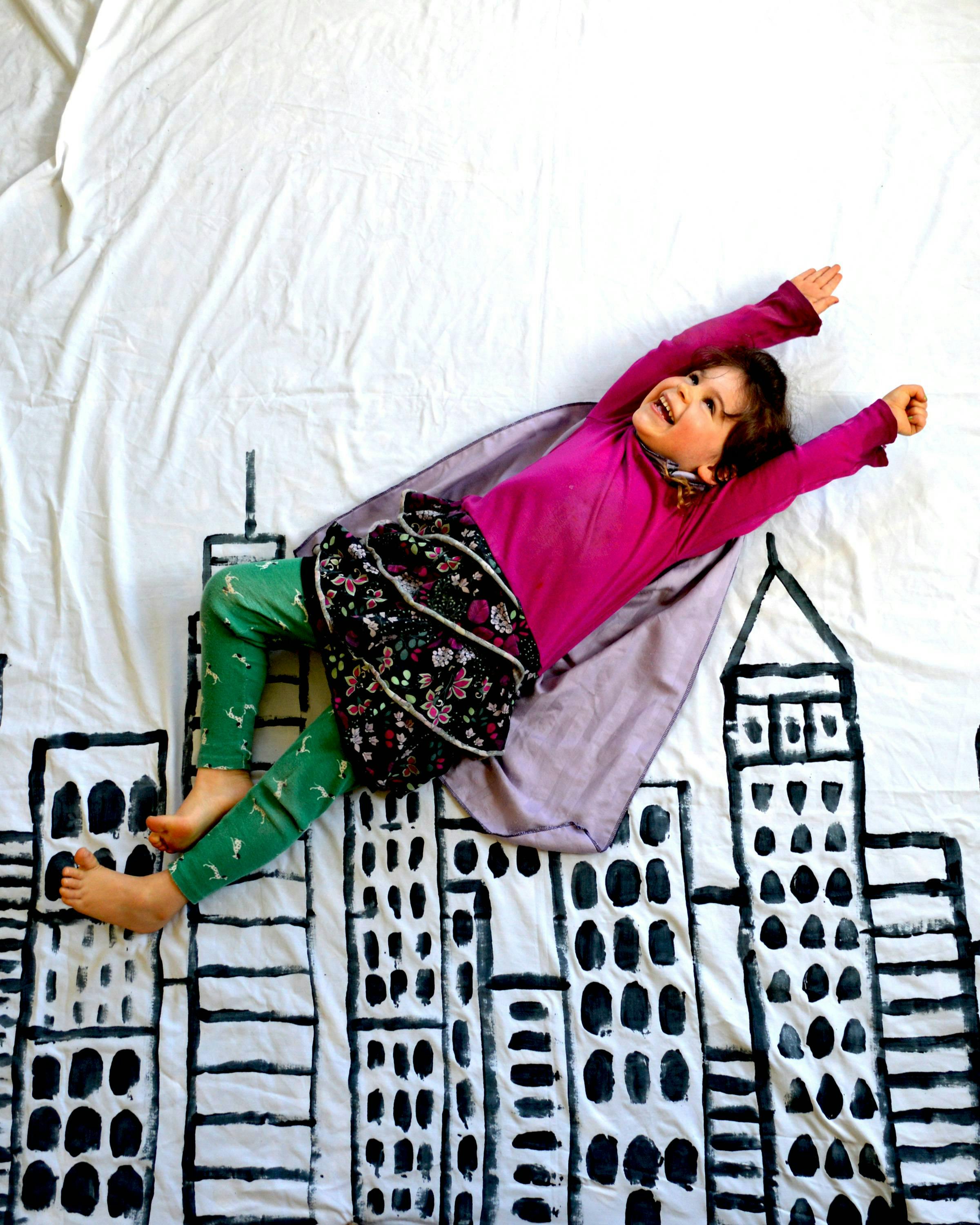 Child in a cape lying on a sheet with drawn buildings