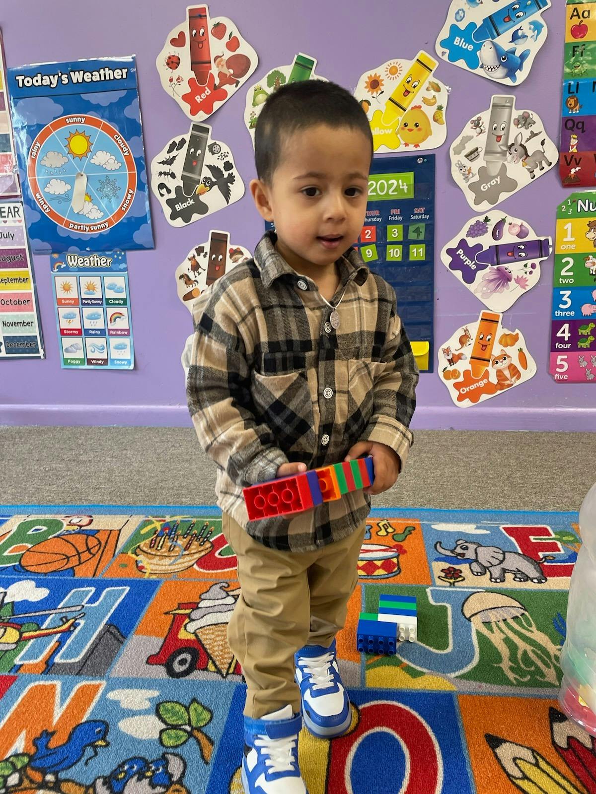 Child standing on a colorful rug holding building blocks in a classroom