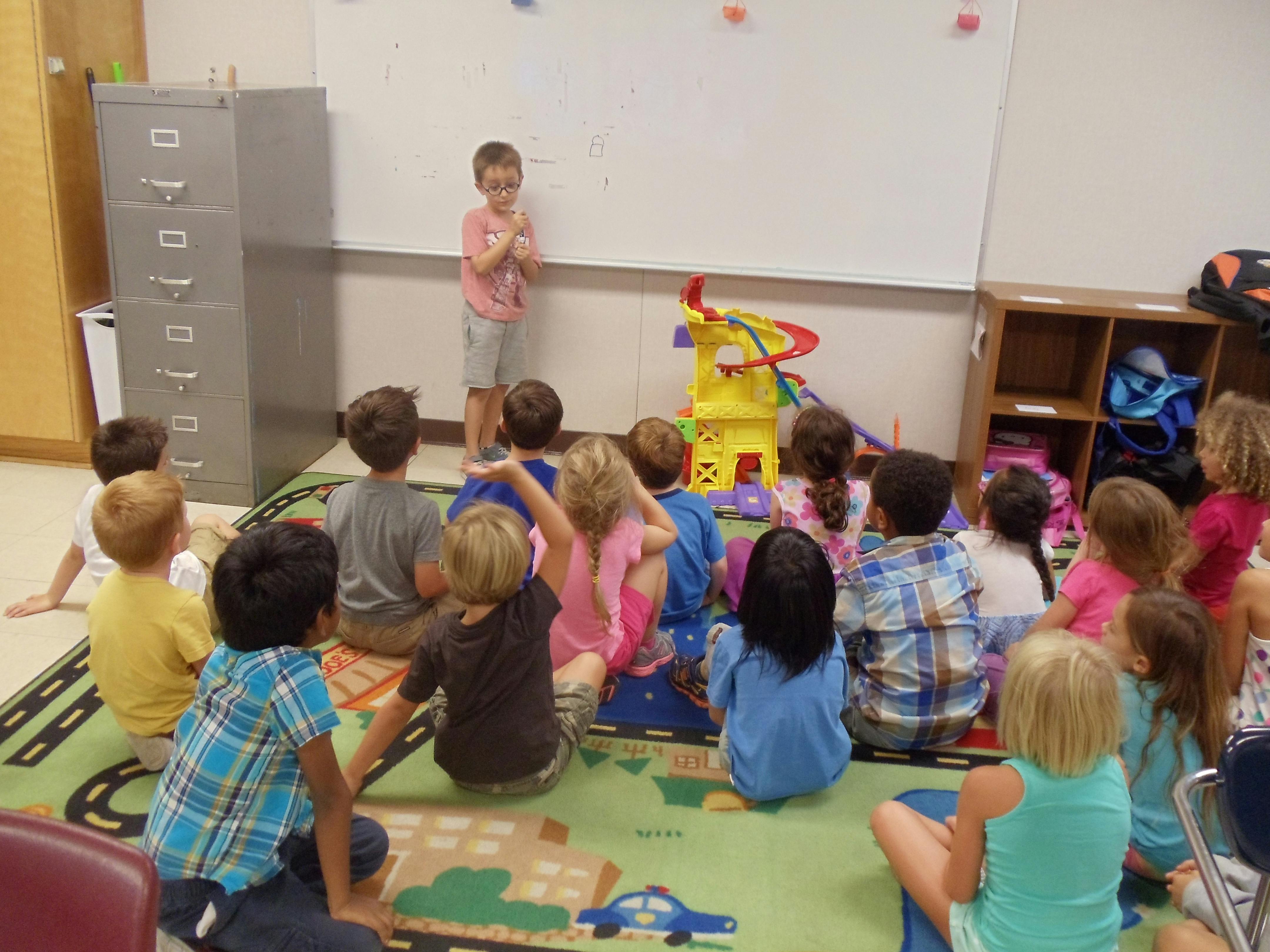 Child standing in front of a whiteboard, speaking to other children seated on a rug
