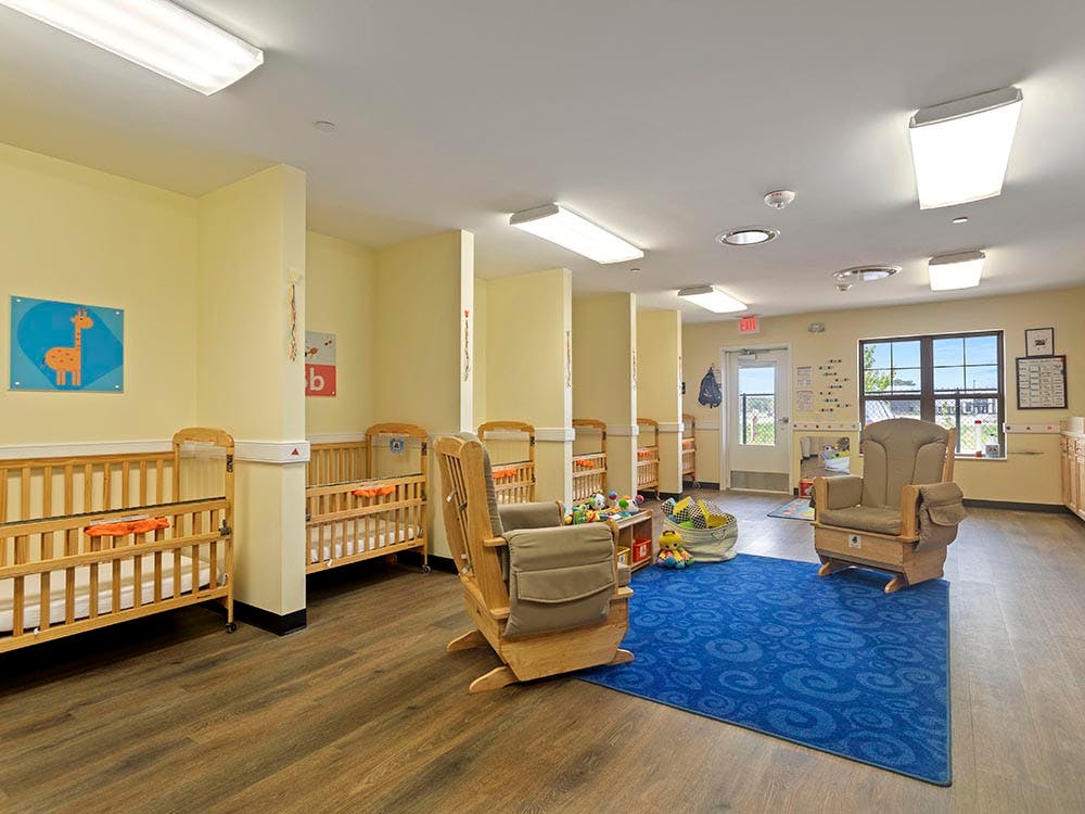 A brightly lit nursery room with several wooden cribs and rocking chairs