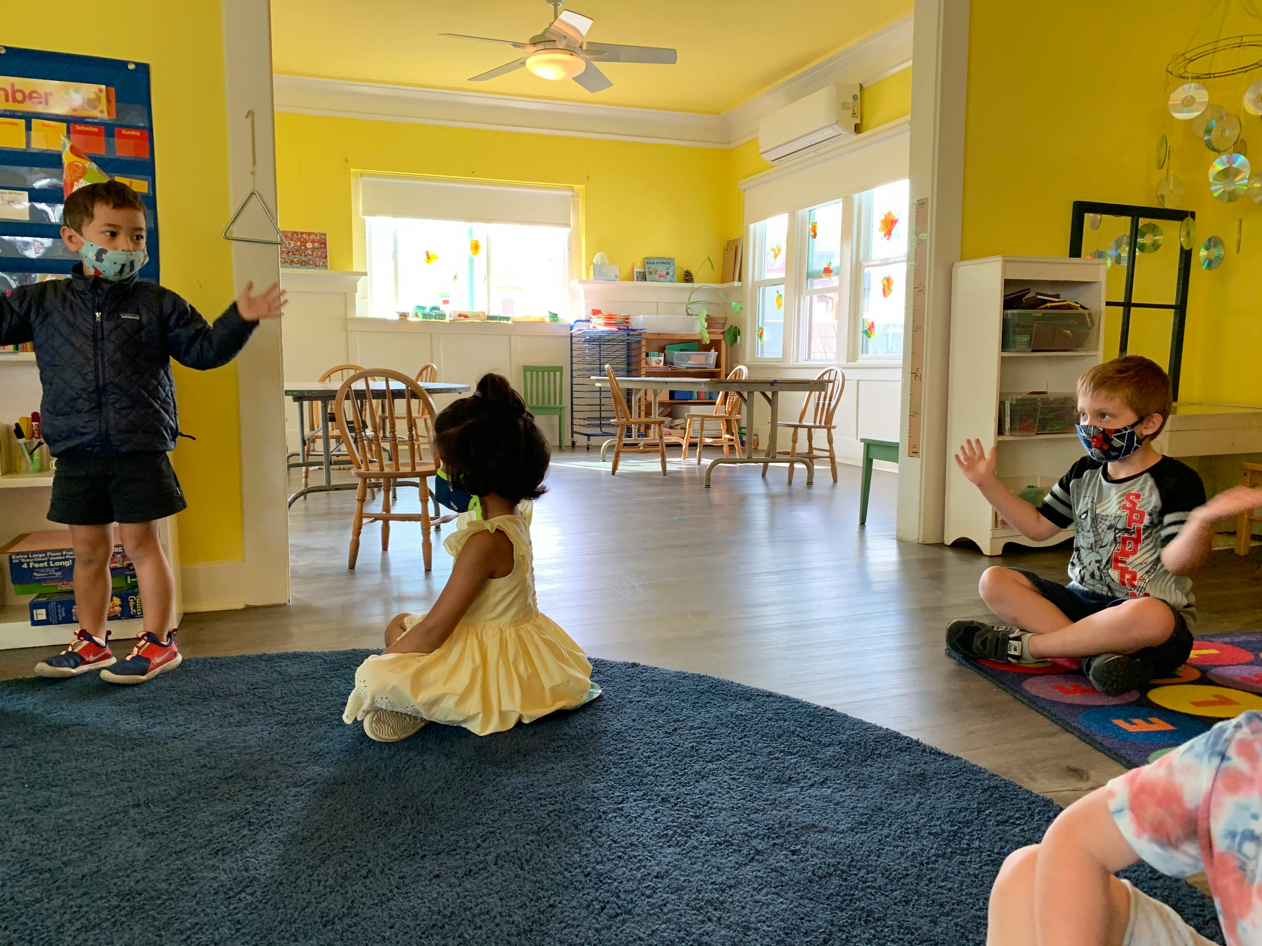 Three children in a classroom, two standing and one sitting on the floor