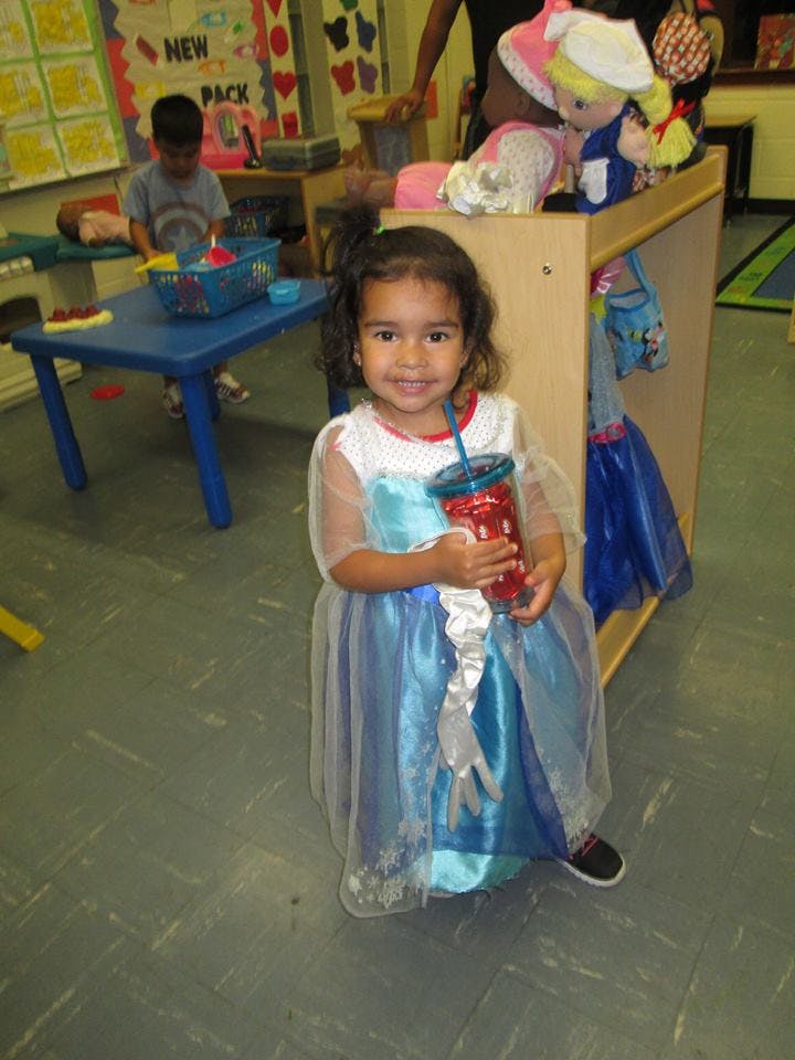 Child in a blue dress holding a red cup with a straw in a classroom