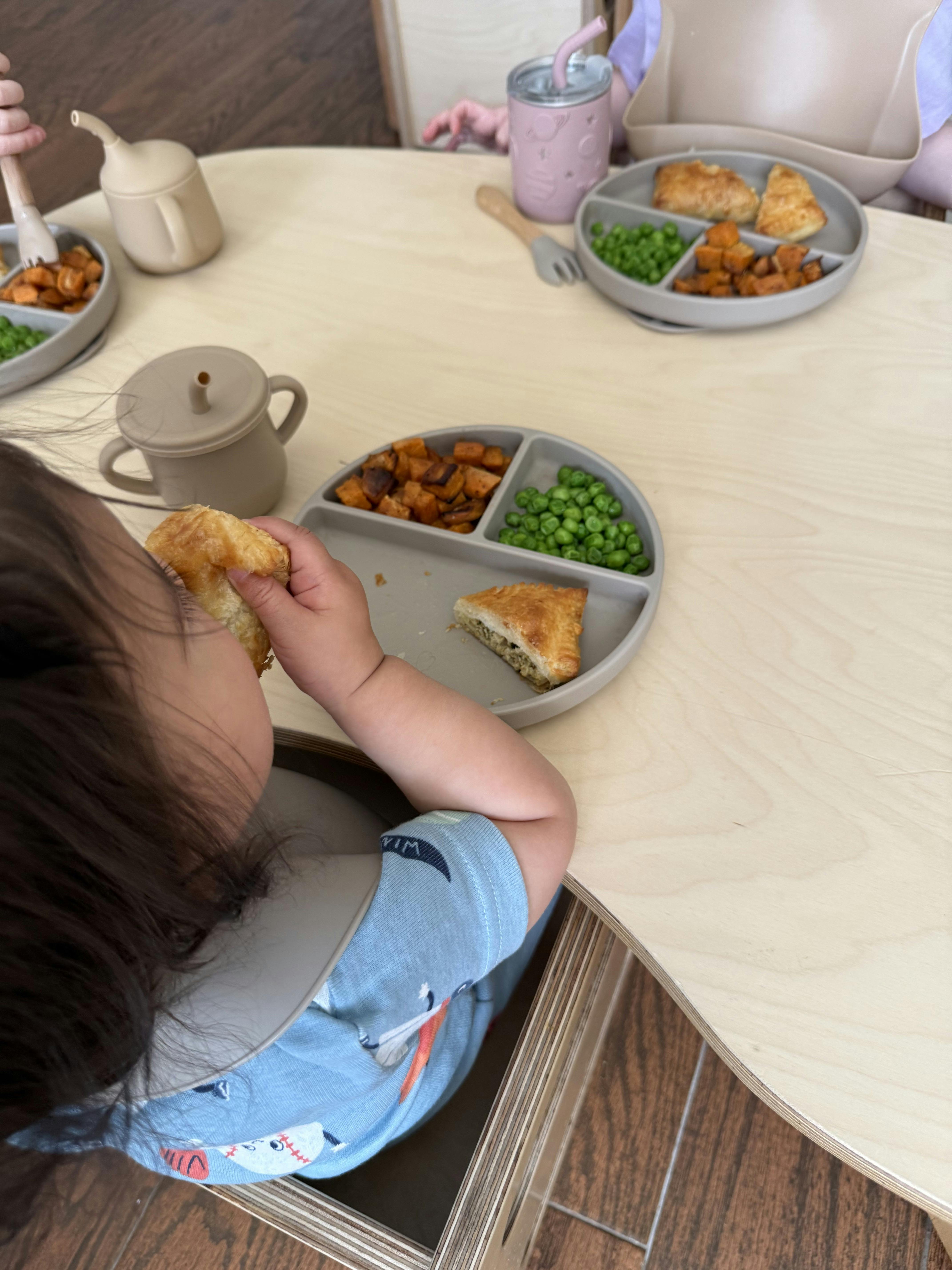 Child in a high chair eating from a plate with peas, sweet potatoes, and pastry