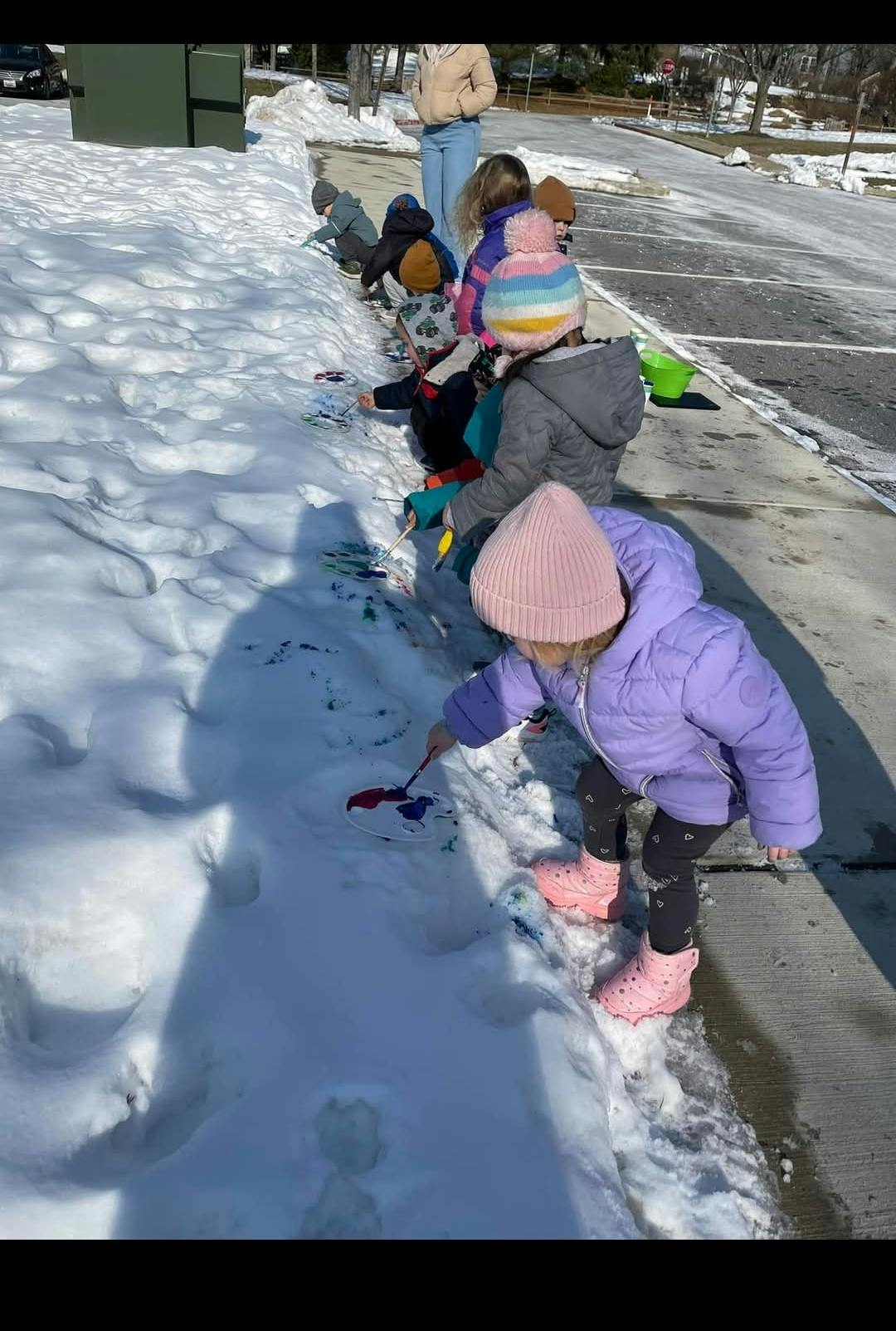Child in purple coat and pink hat painting snow with a brush in the snow