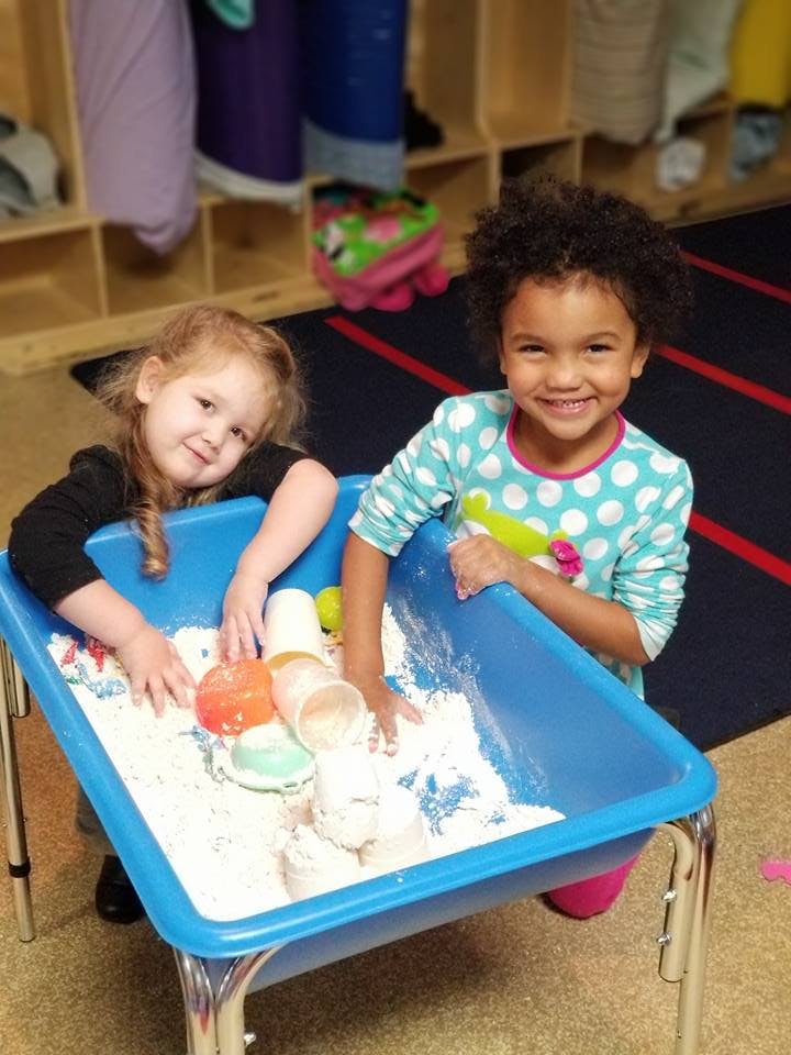 Two children playing in a sensory bin with white sand and colorful objects