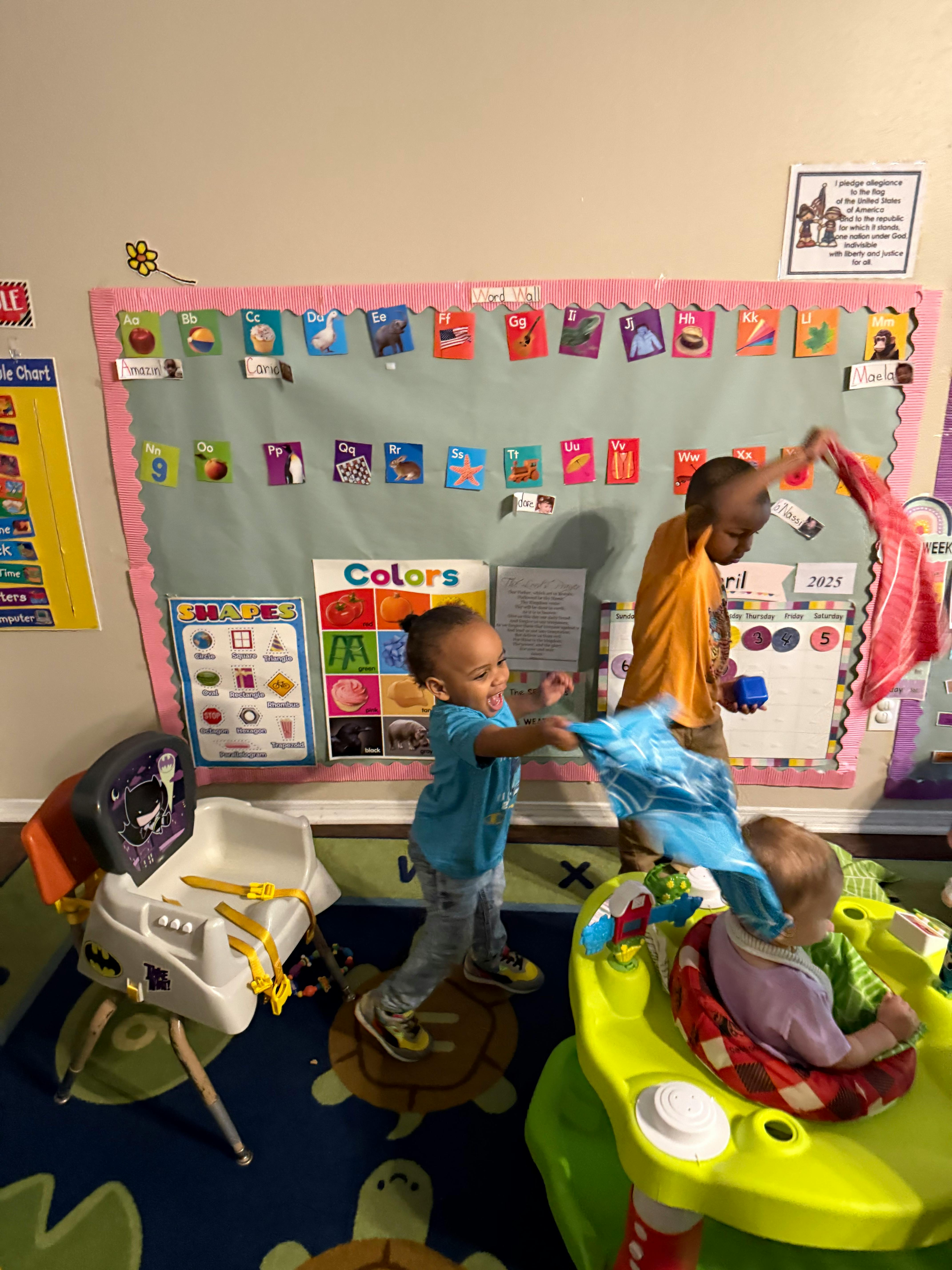 Two children playing with fabric in a classroom, one child in a baby seat