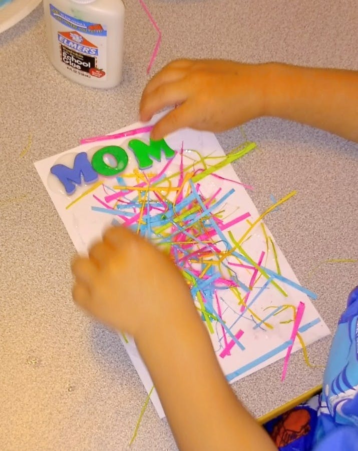 Child's hands creating a card with the word "MOM" and colorful strips of paper