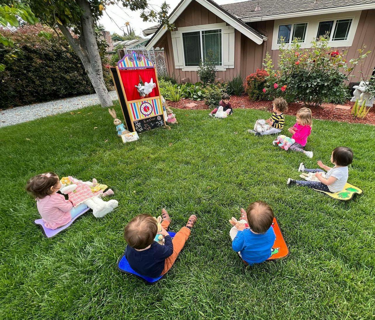 Children sitting on grass in a circle facing a puppet show stage