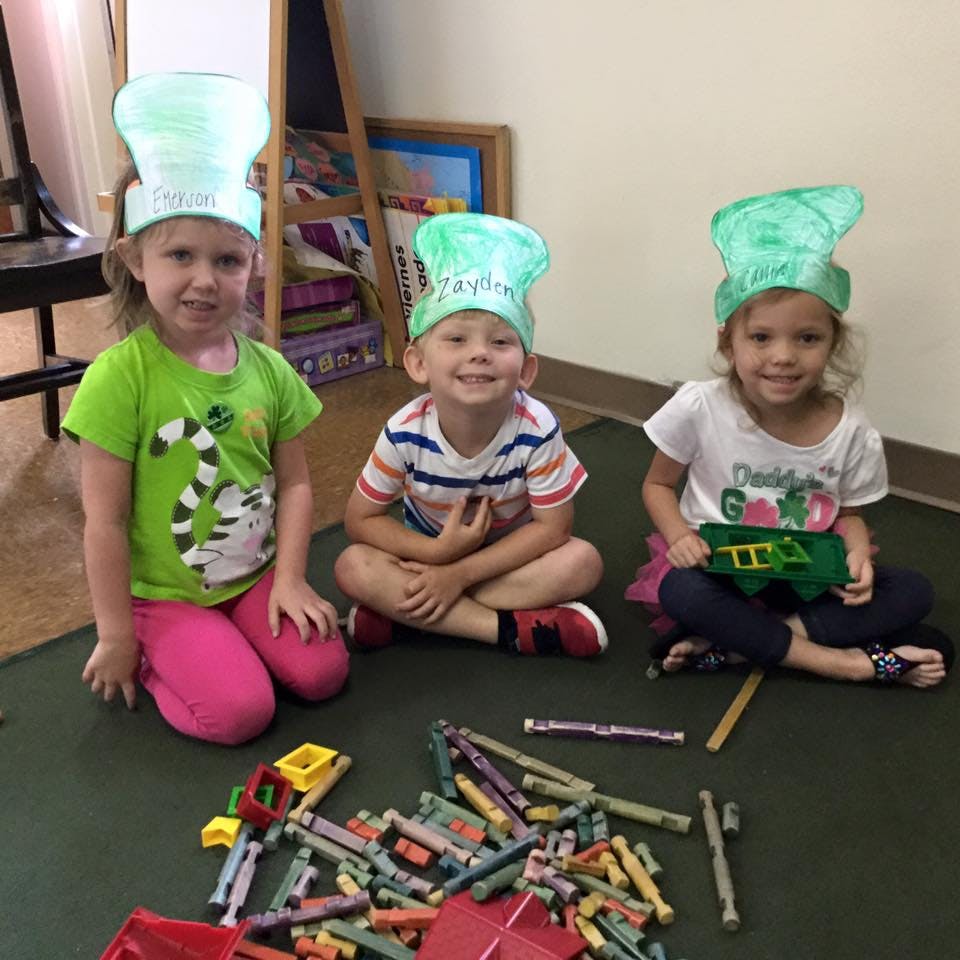 Three children wearing green paper hats sitting on the floor with toys
