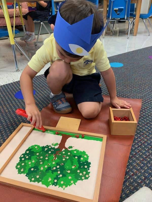 Child in a paper crown placing small red objects on a tree drawing