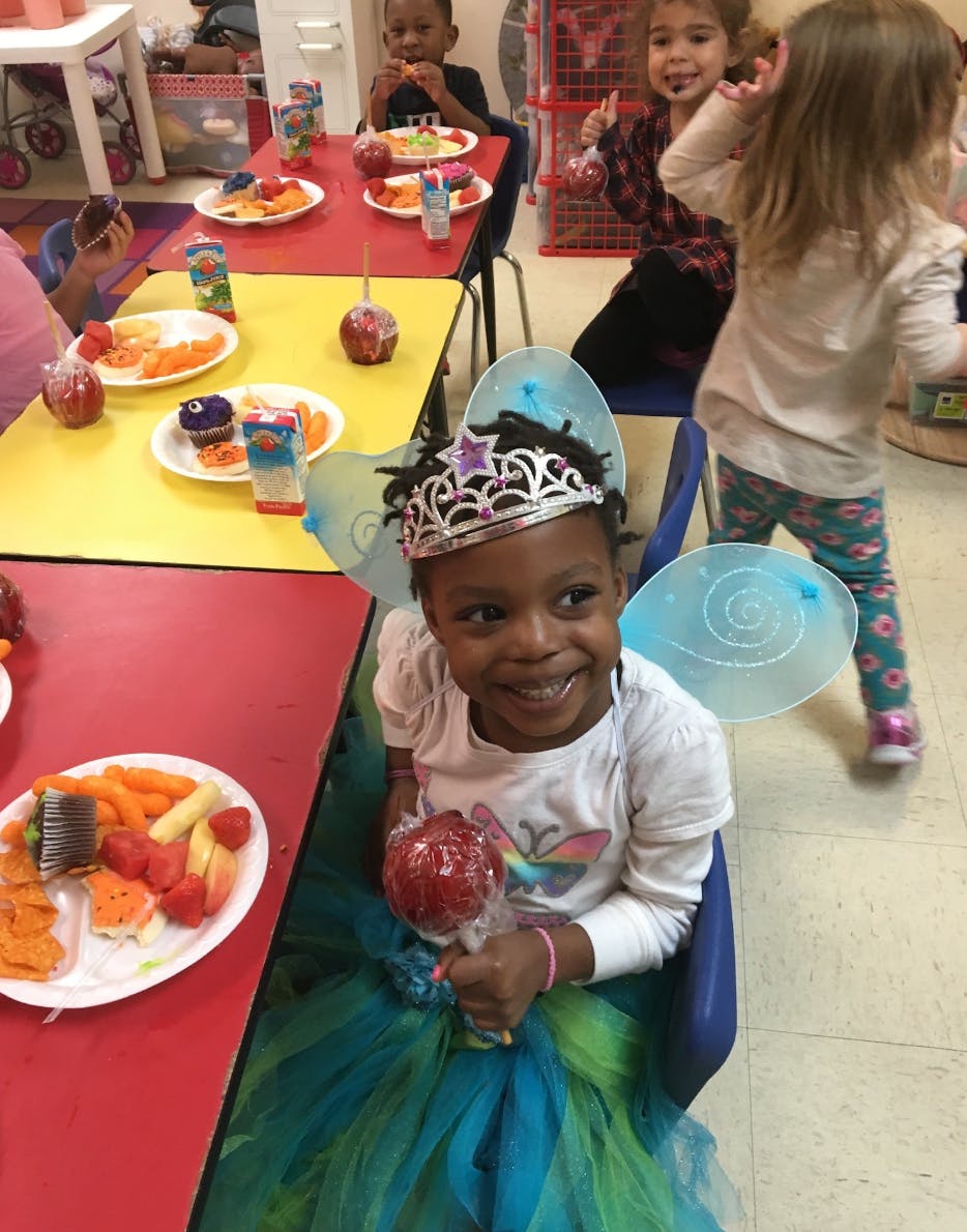 Child in a crown and wings holding a candy apple at a table with other children