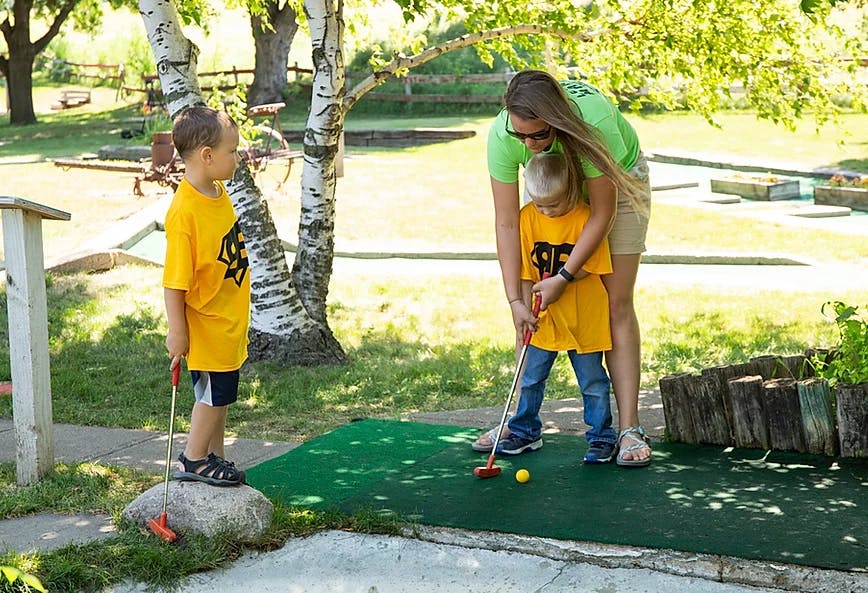 Person helping a child play mini golf while another child watches