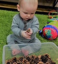 Child sitting on a green rug holding a pinecone over a container of pinecones