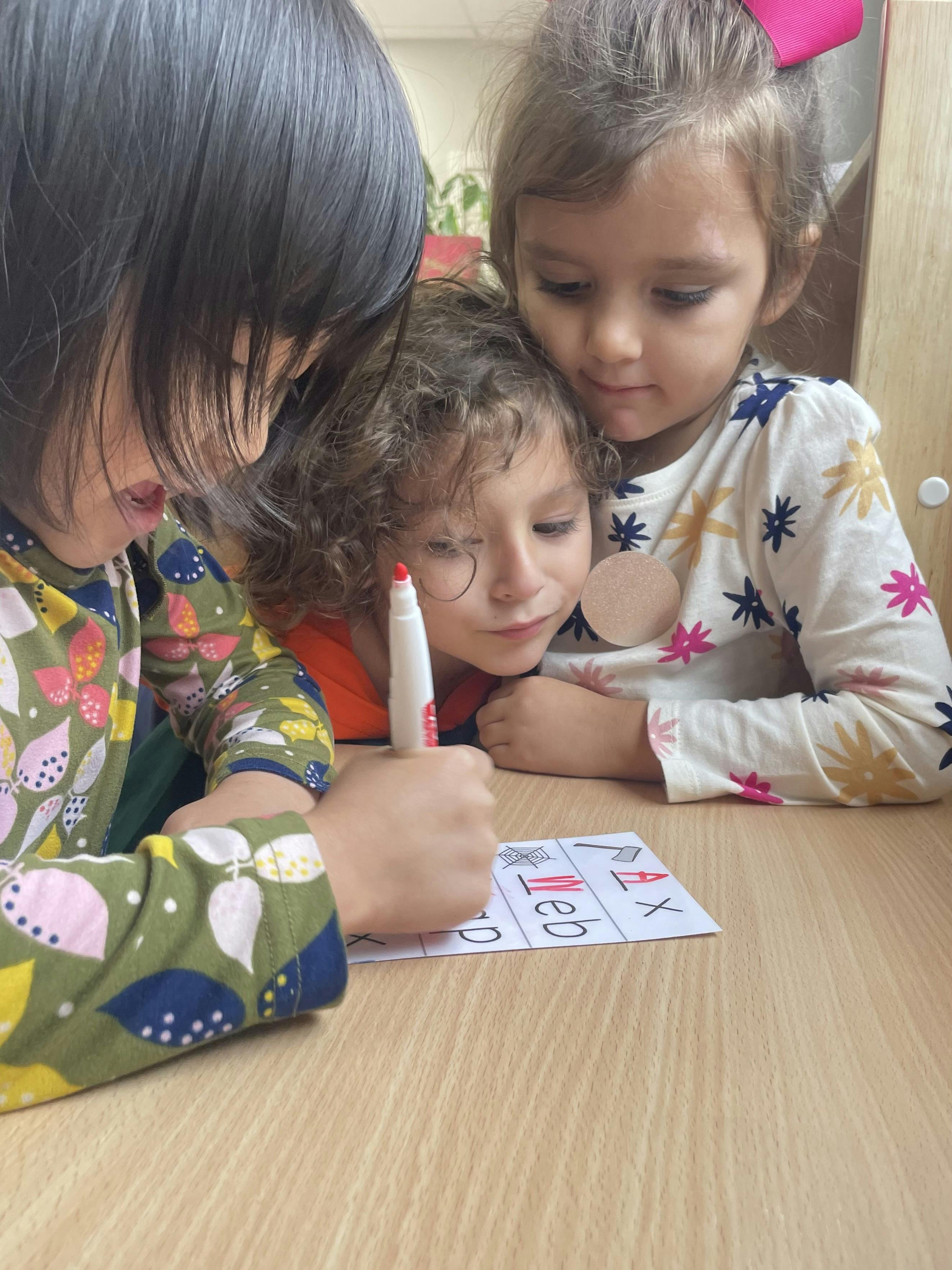 Three children looking at a paper with letters, one child holding a marker