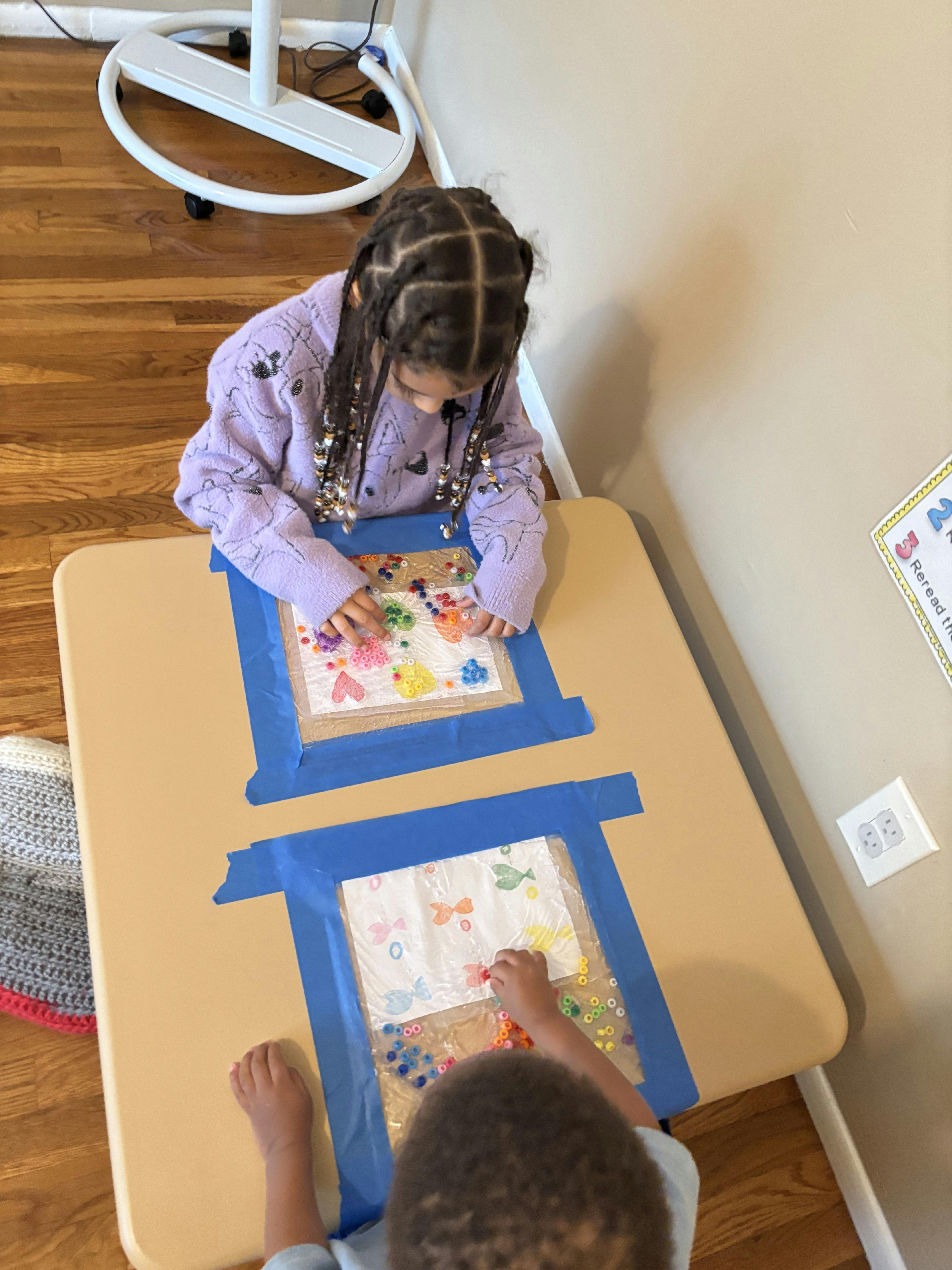 Two children arranging beads on adhesive sheets taped to a table