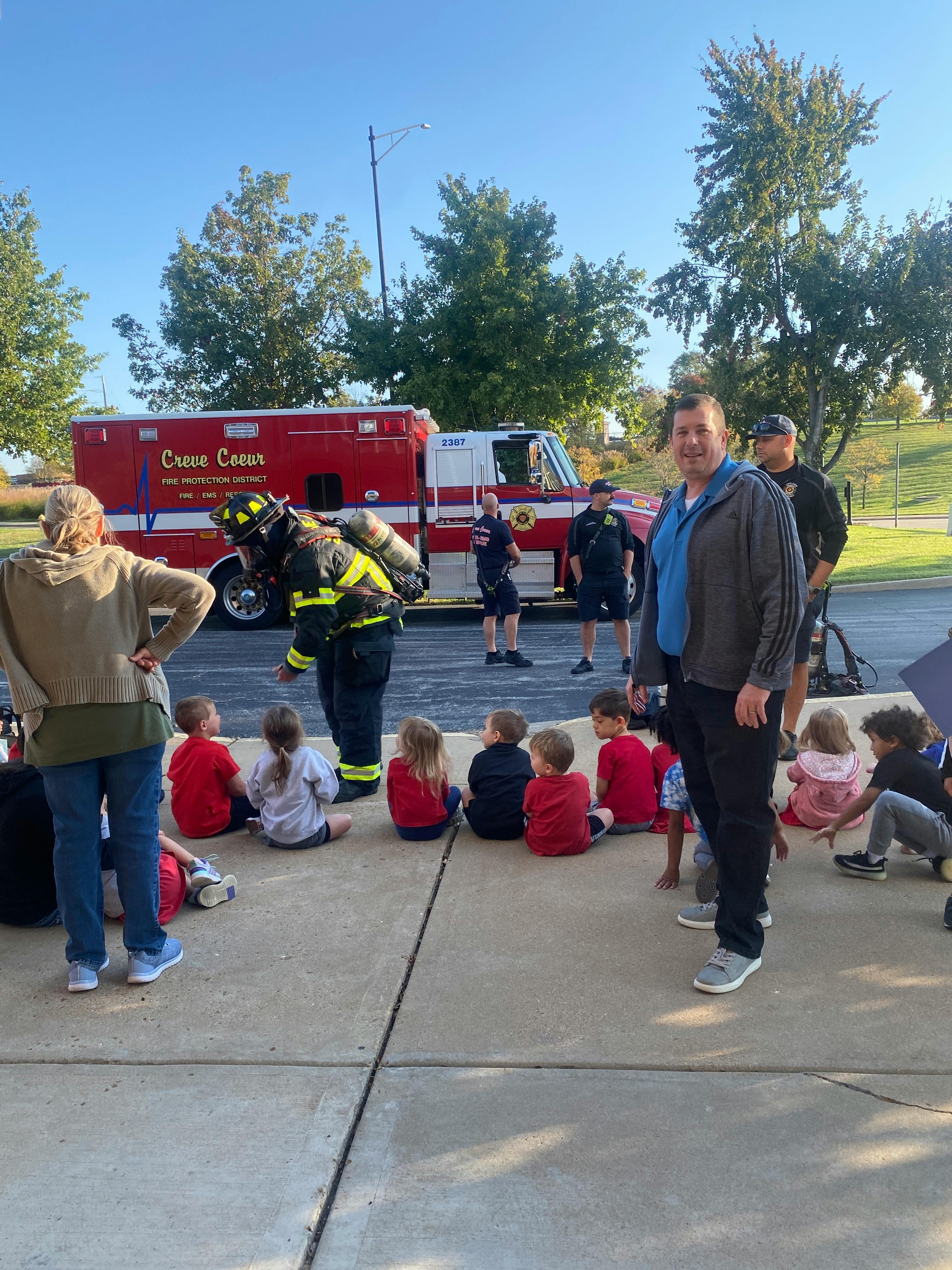 Firefighter in gear talking to children sitting on the ground near a fire truck