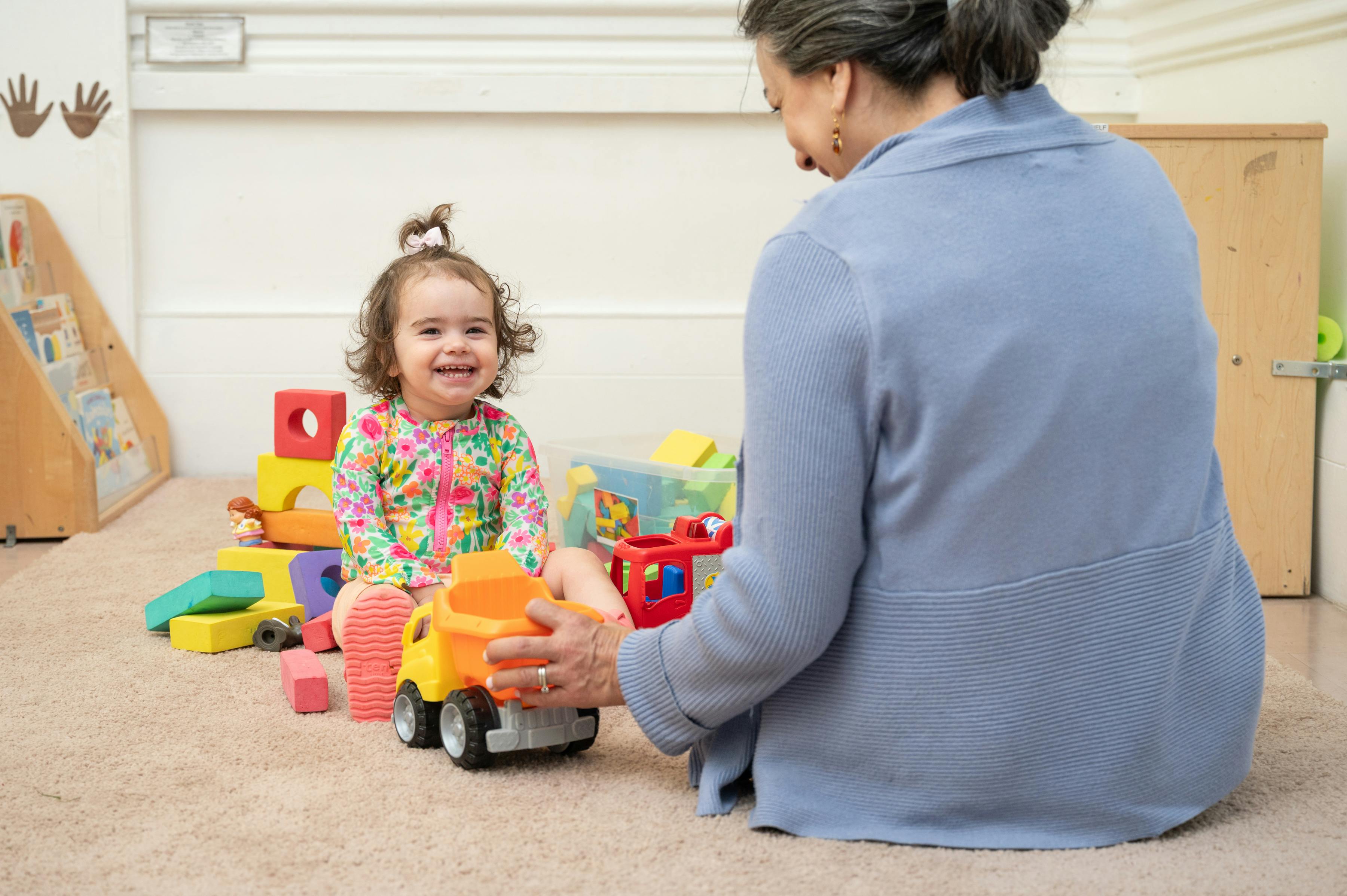 A person and a child sitting on a floor playing with toy trucks and blocks