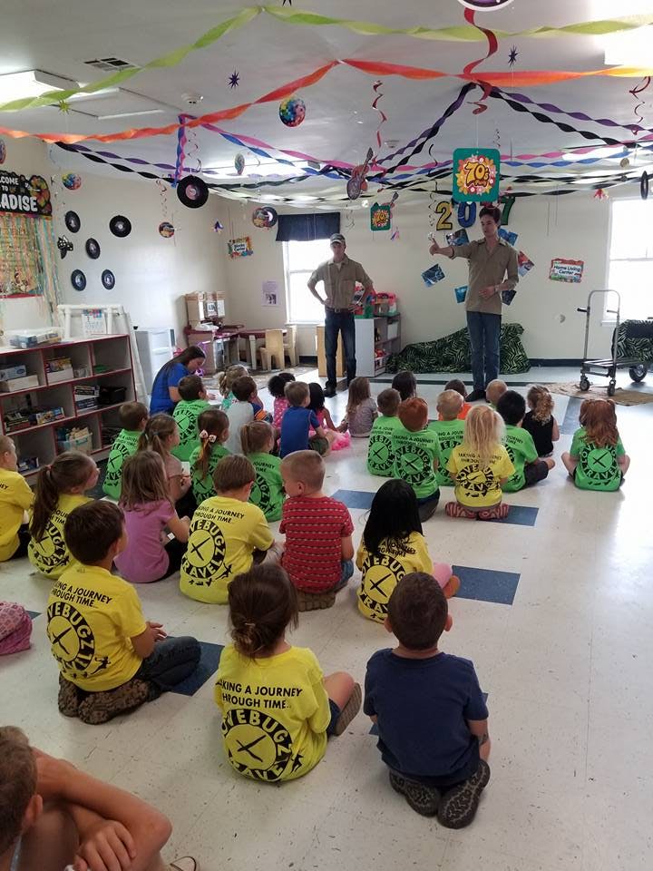 Two people standing in front of a group of children sitting on the floor in a classroom