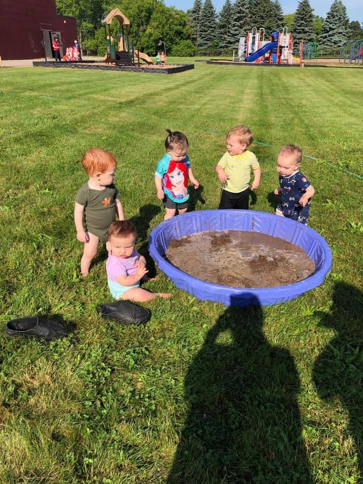 Children playing around a small pool filled with mud on a grassy lawn