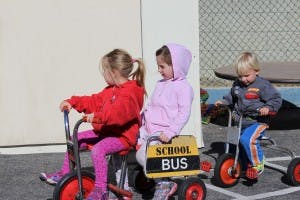 Three children riding tricycles, one with a "SCHOOL BUS" sign