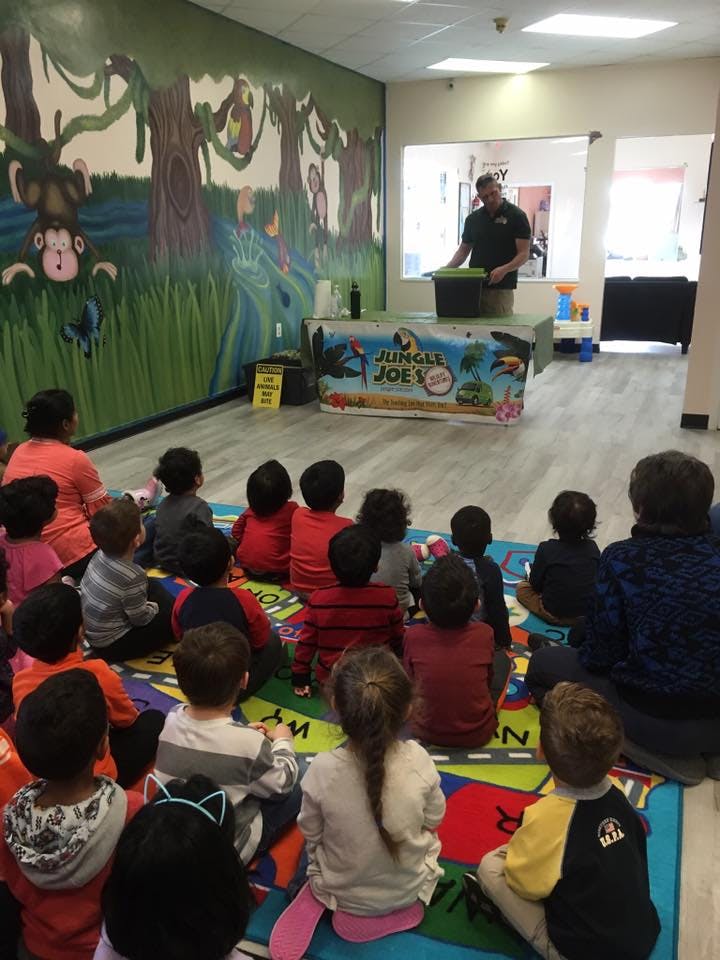 Children sitting on a rug watching a person standing behind a table