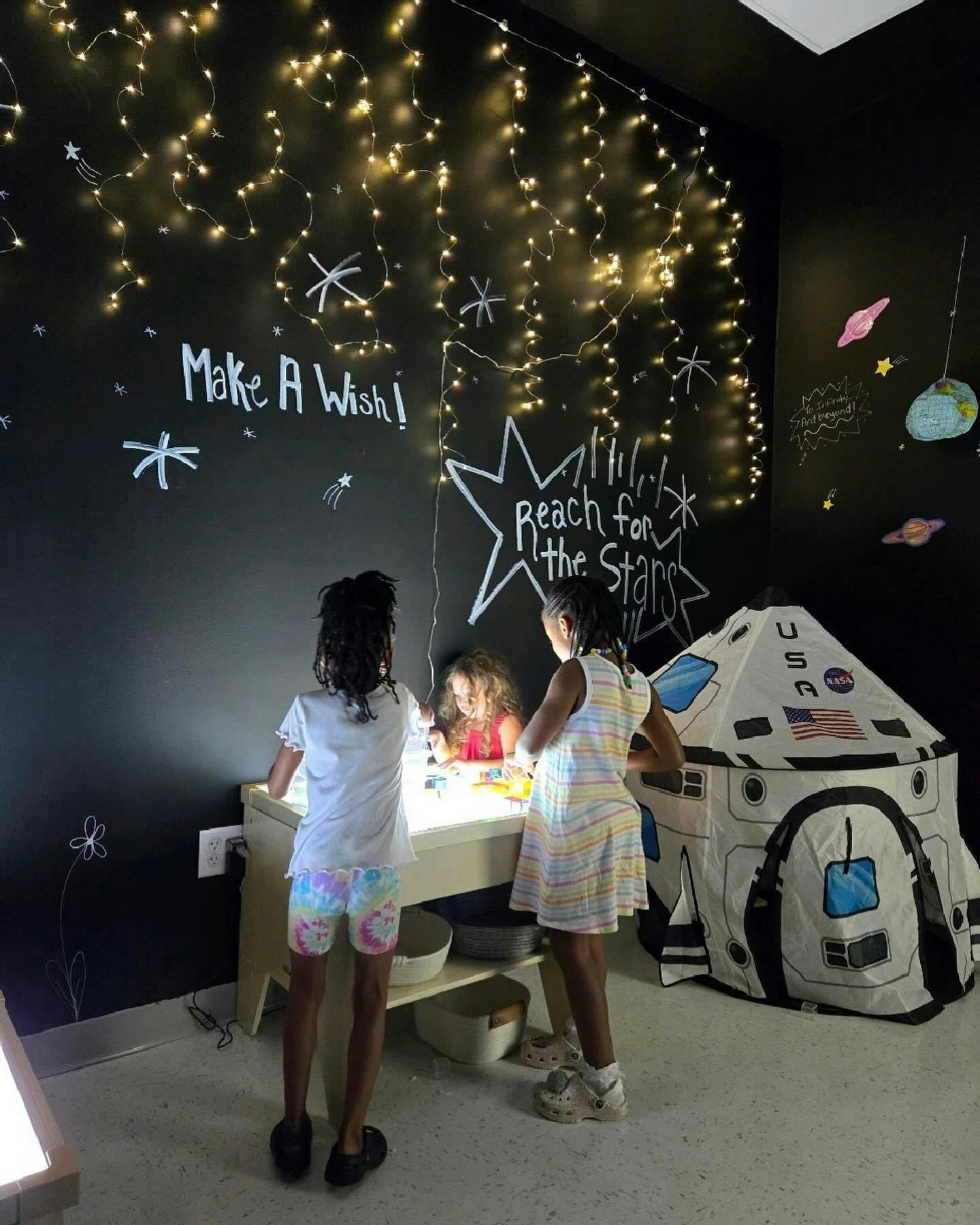 Three children playing at a light table in a dark room with "Make A Wish" written on the wall