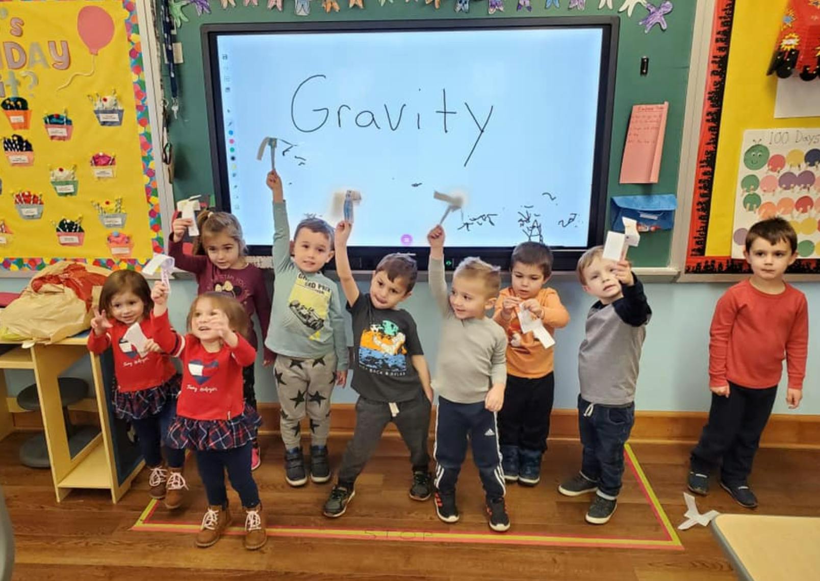 Children holding up paper airplanes in front of a screen displaying "Gravity"