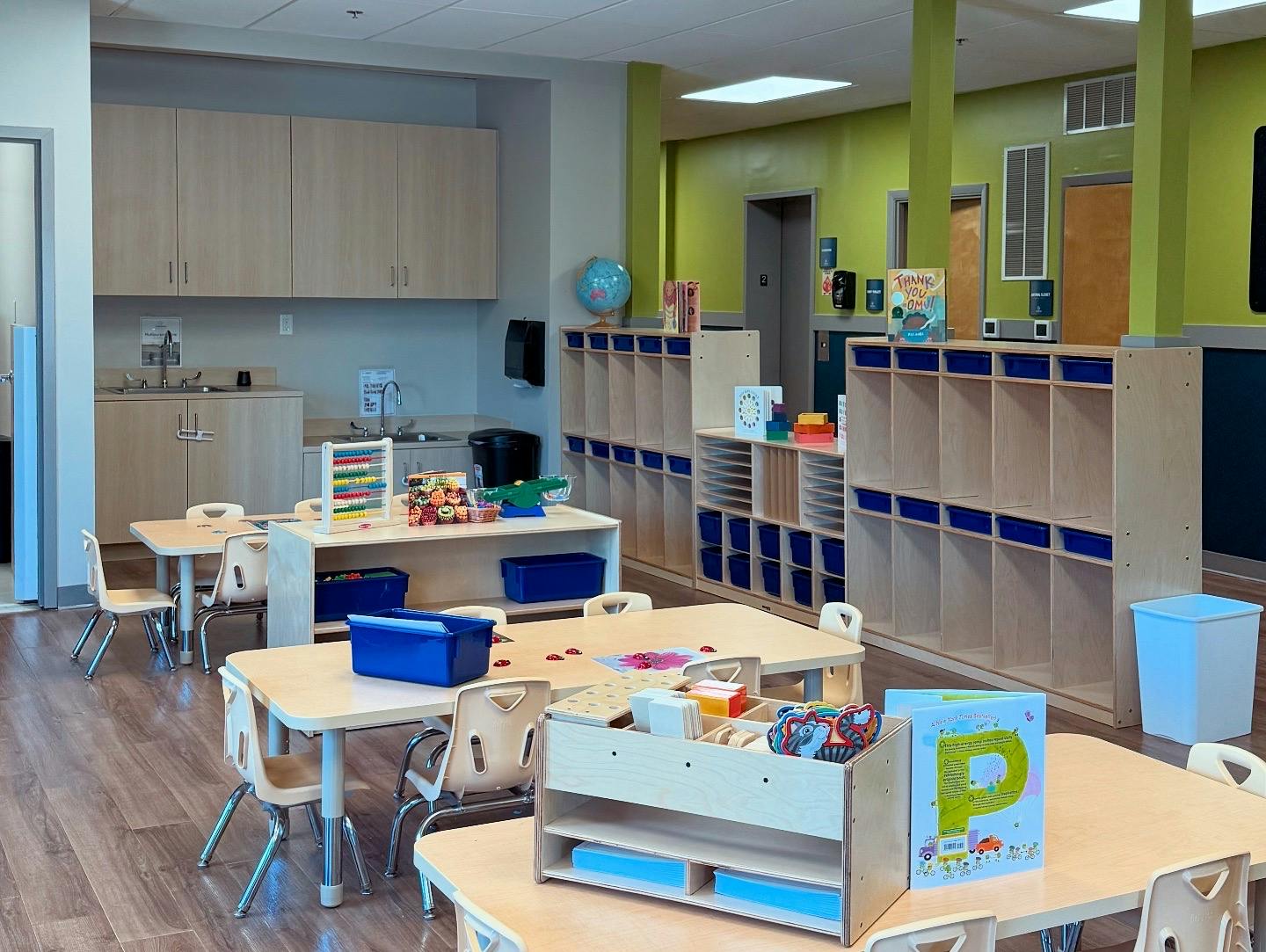 Empty classroom with small tables and chairs, storage cubbies, and a sink area