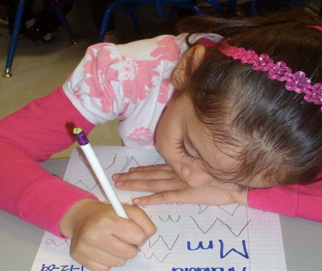 Child writing letters on paper with a pen, head resting on table