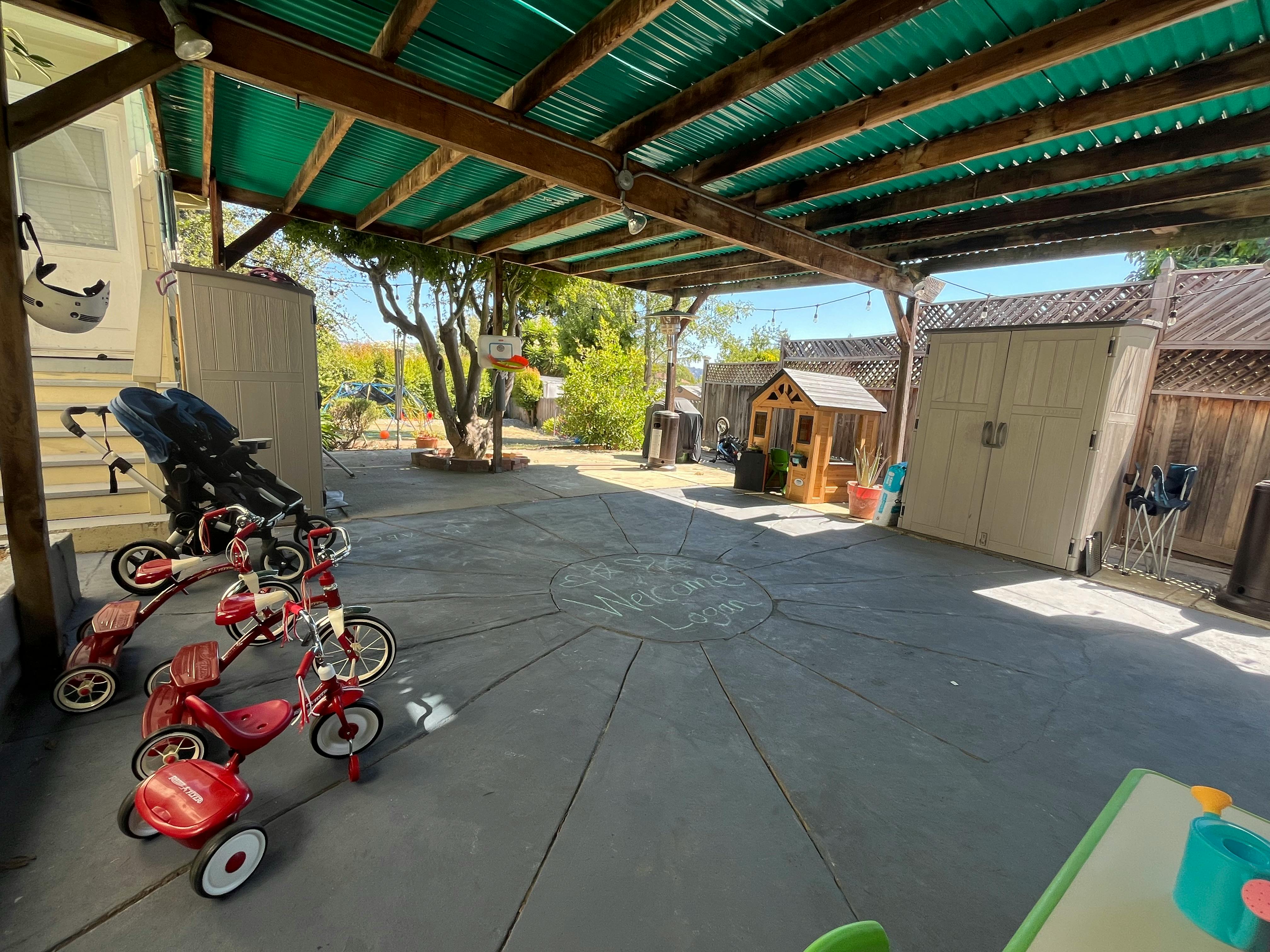 Tricycles and stroller under a covered patio with a playhouse in the background