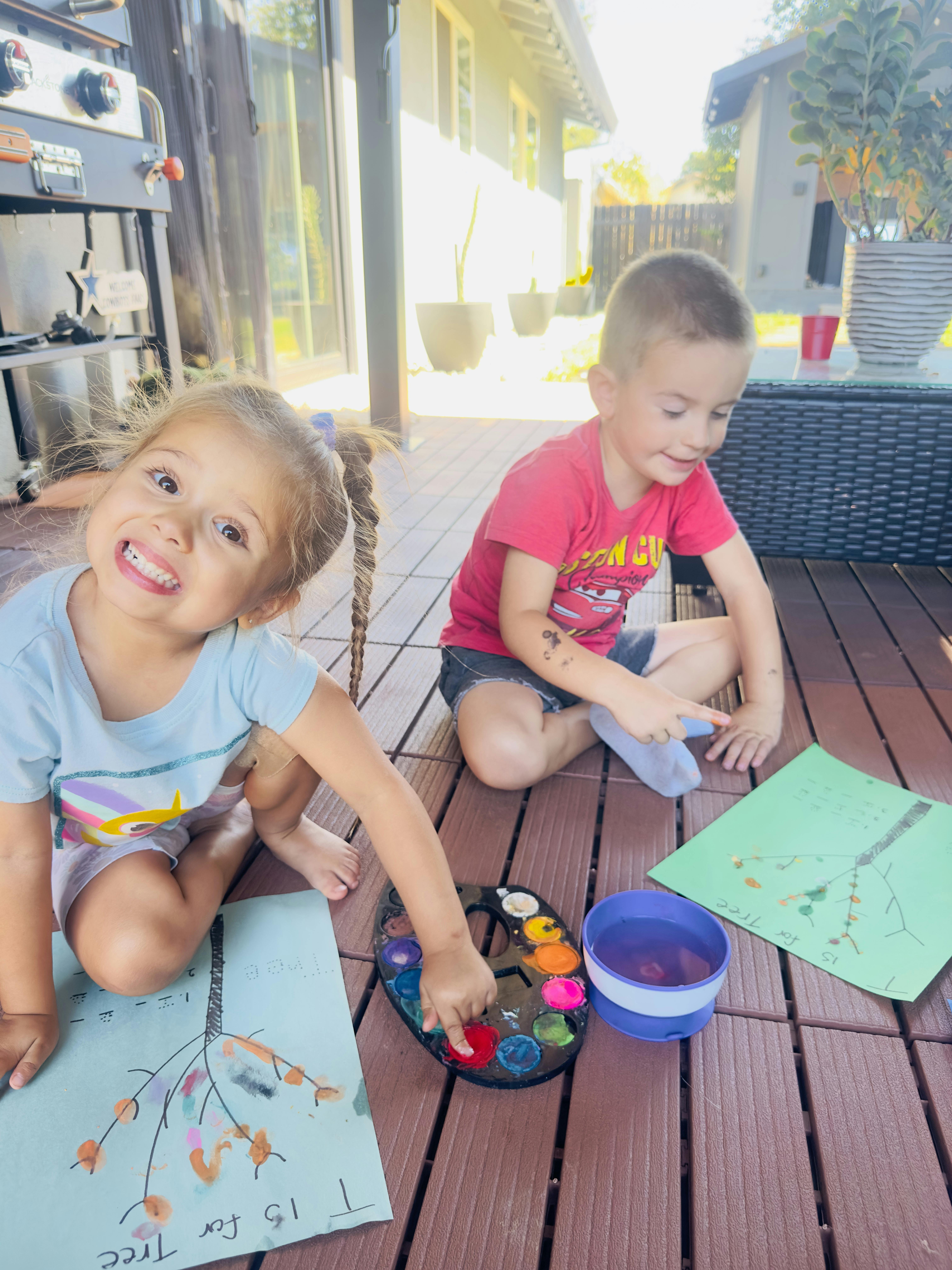Two children painting on paper while sitting on an outdoor deck