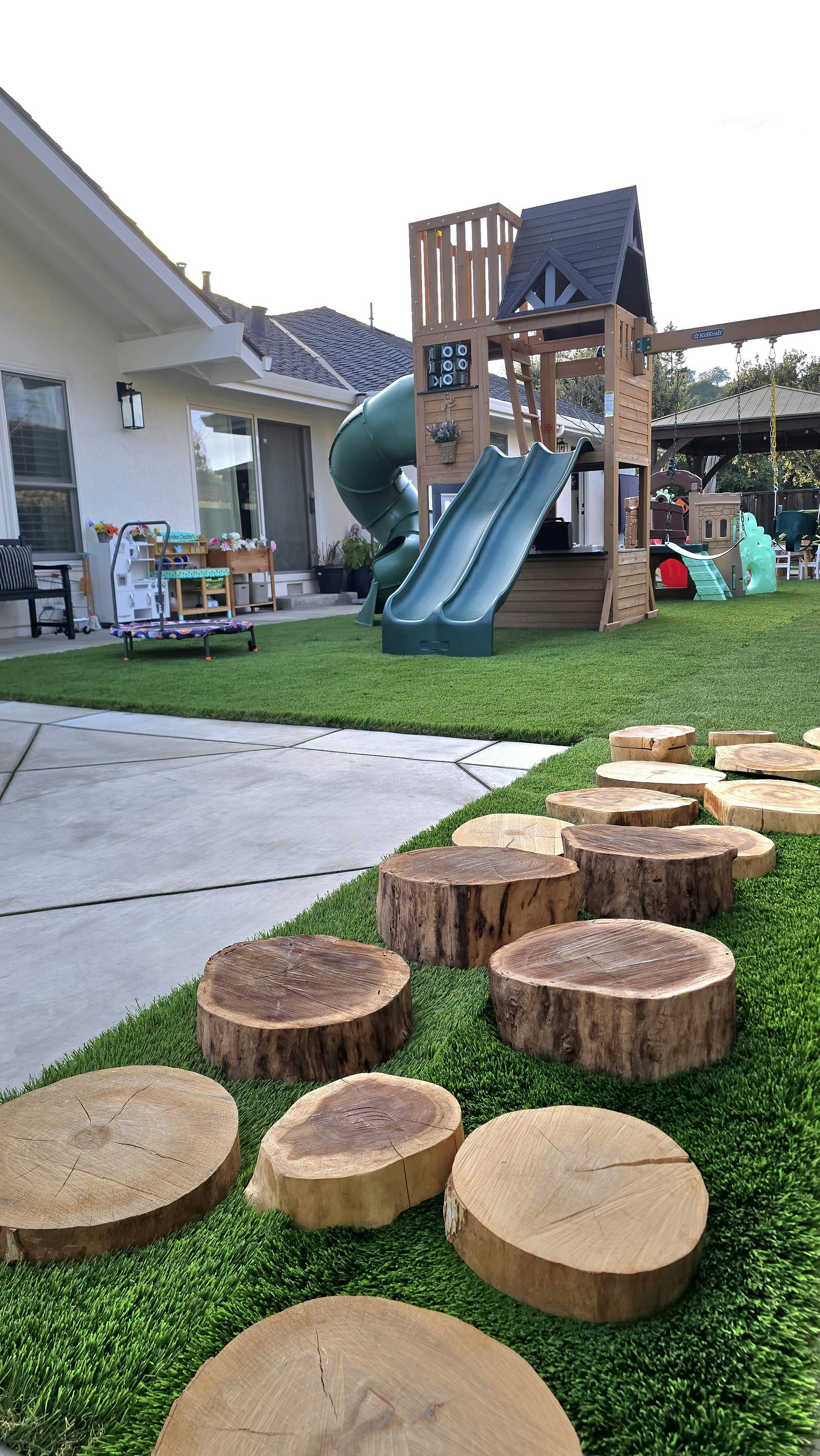 Backyard with an artificial turf lawn, a playset, and tree stump stepping stones