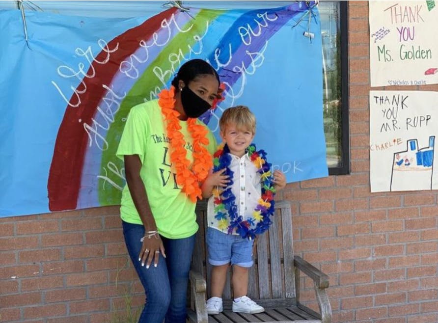 Person in face mask and child in leis standing in front of rainbow banner