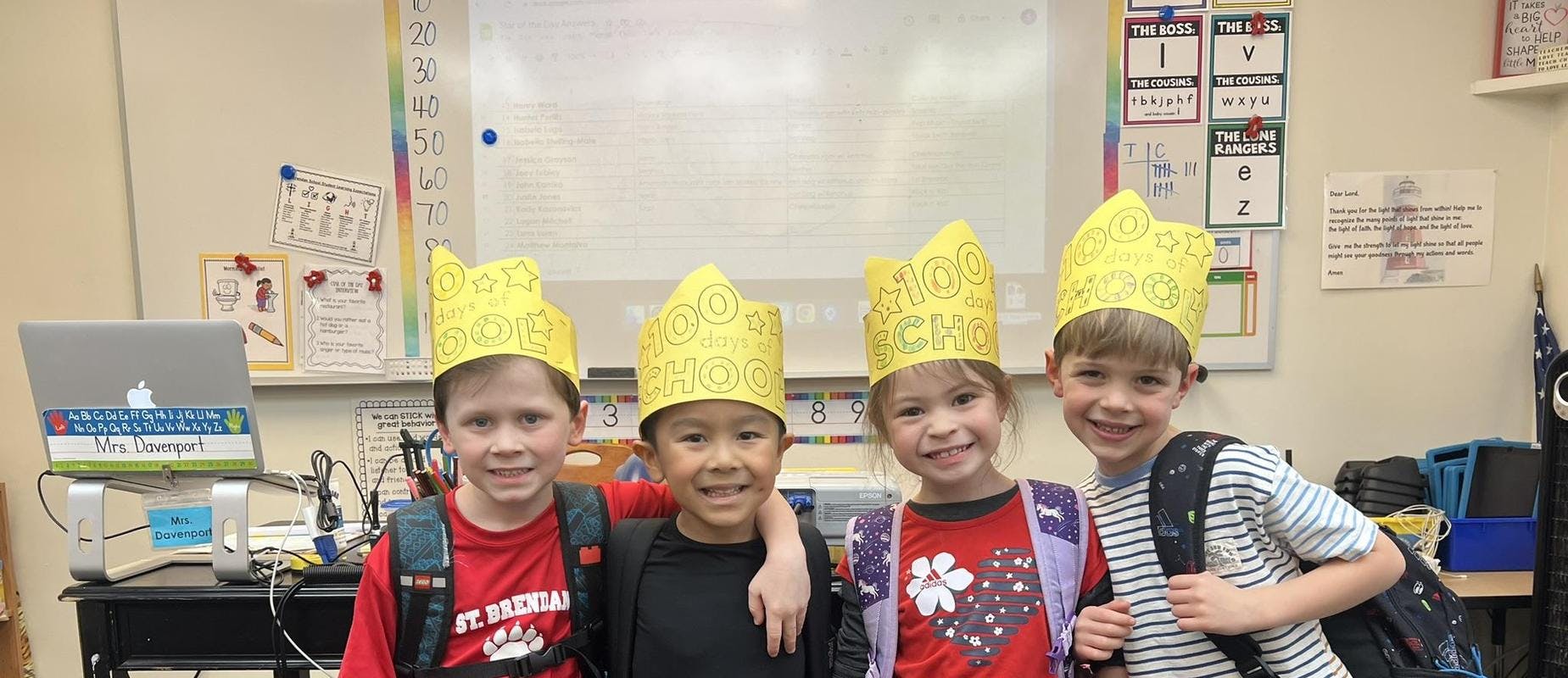 Four children in a classroom wearing yellow "100 days of school" crowns