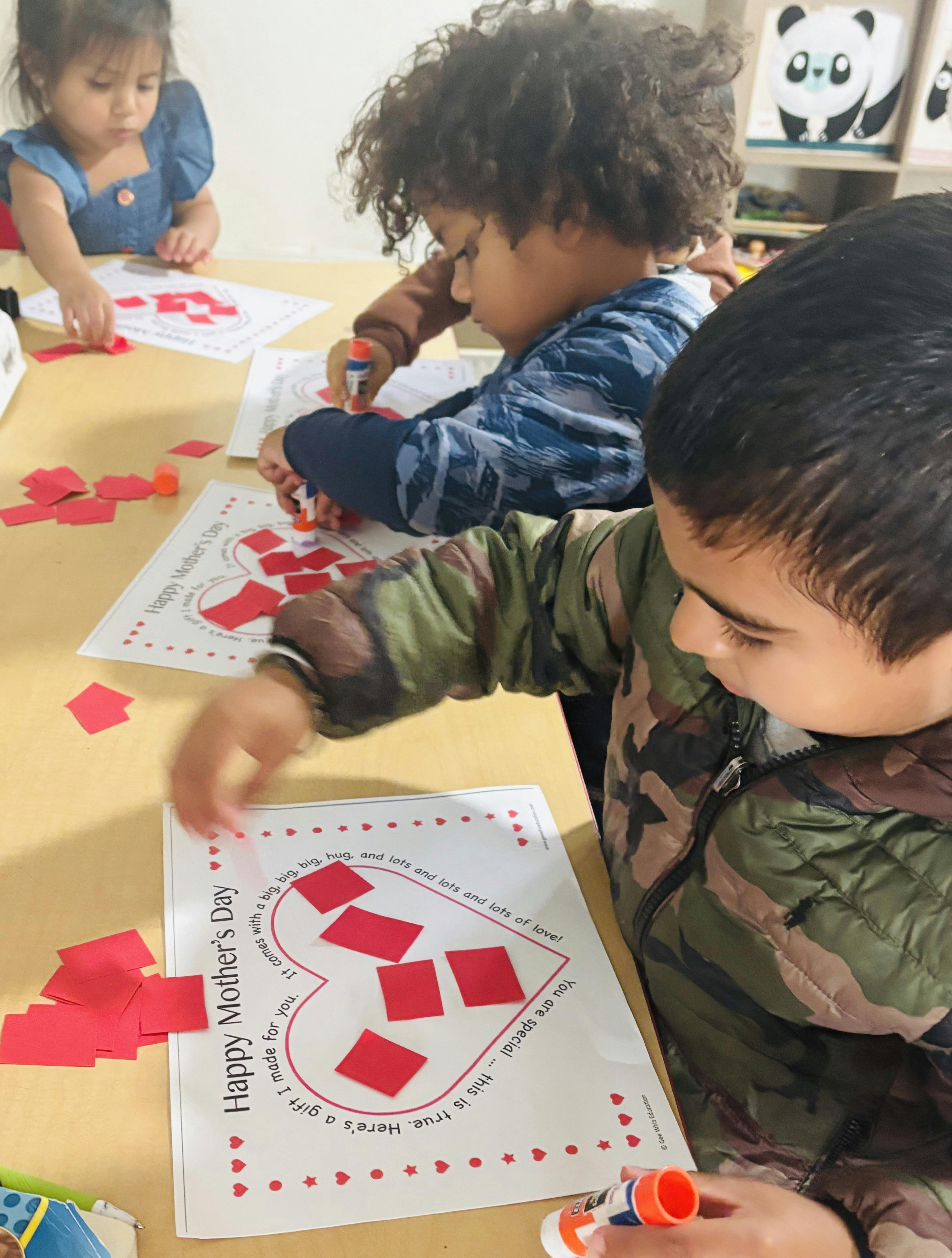 Children gluing red squares onto paper heart shapes that read "Happy Mother's Day"