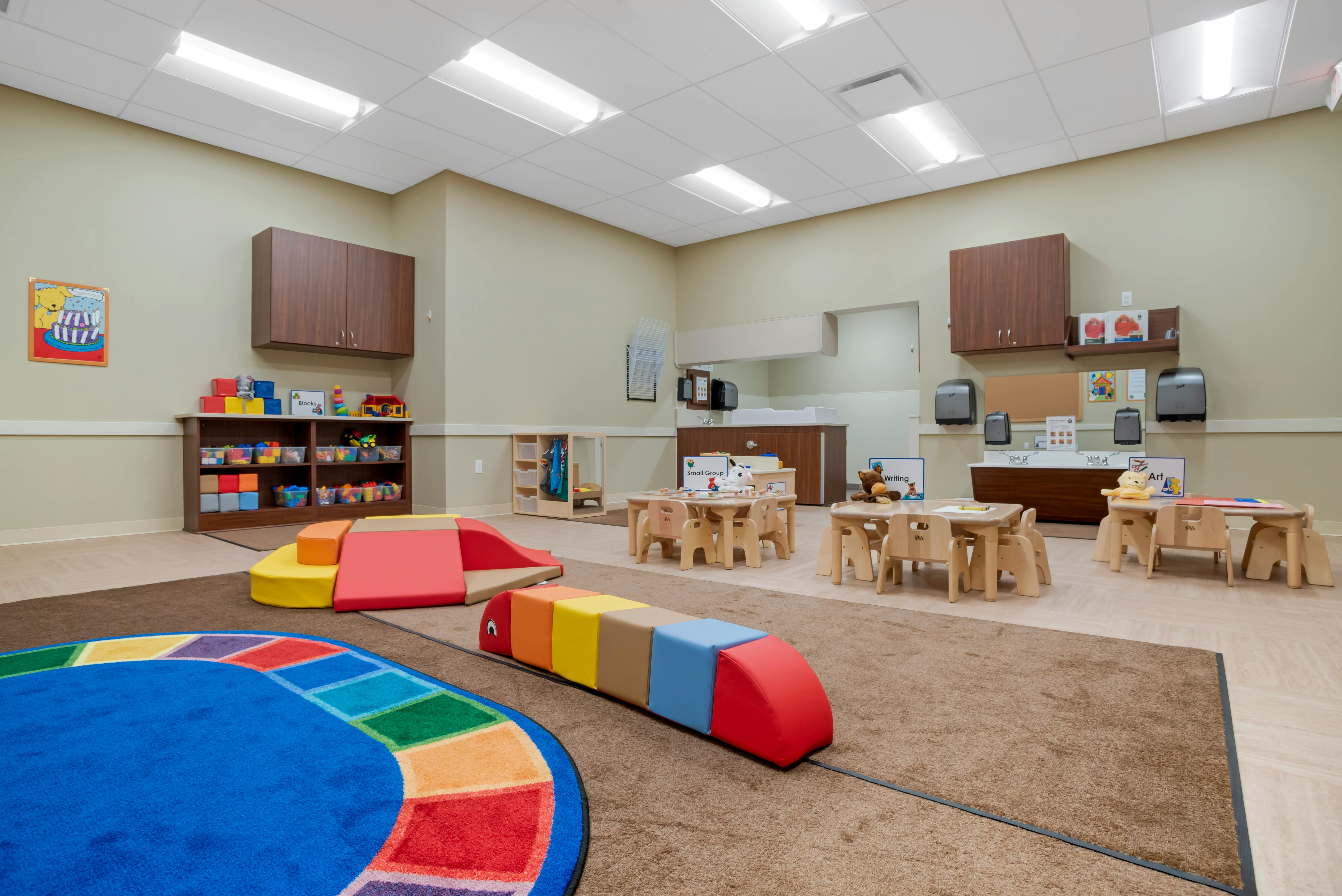 Classroom with small tables and chairs, play mats, and storage shelves with toys