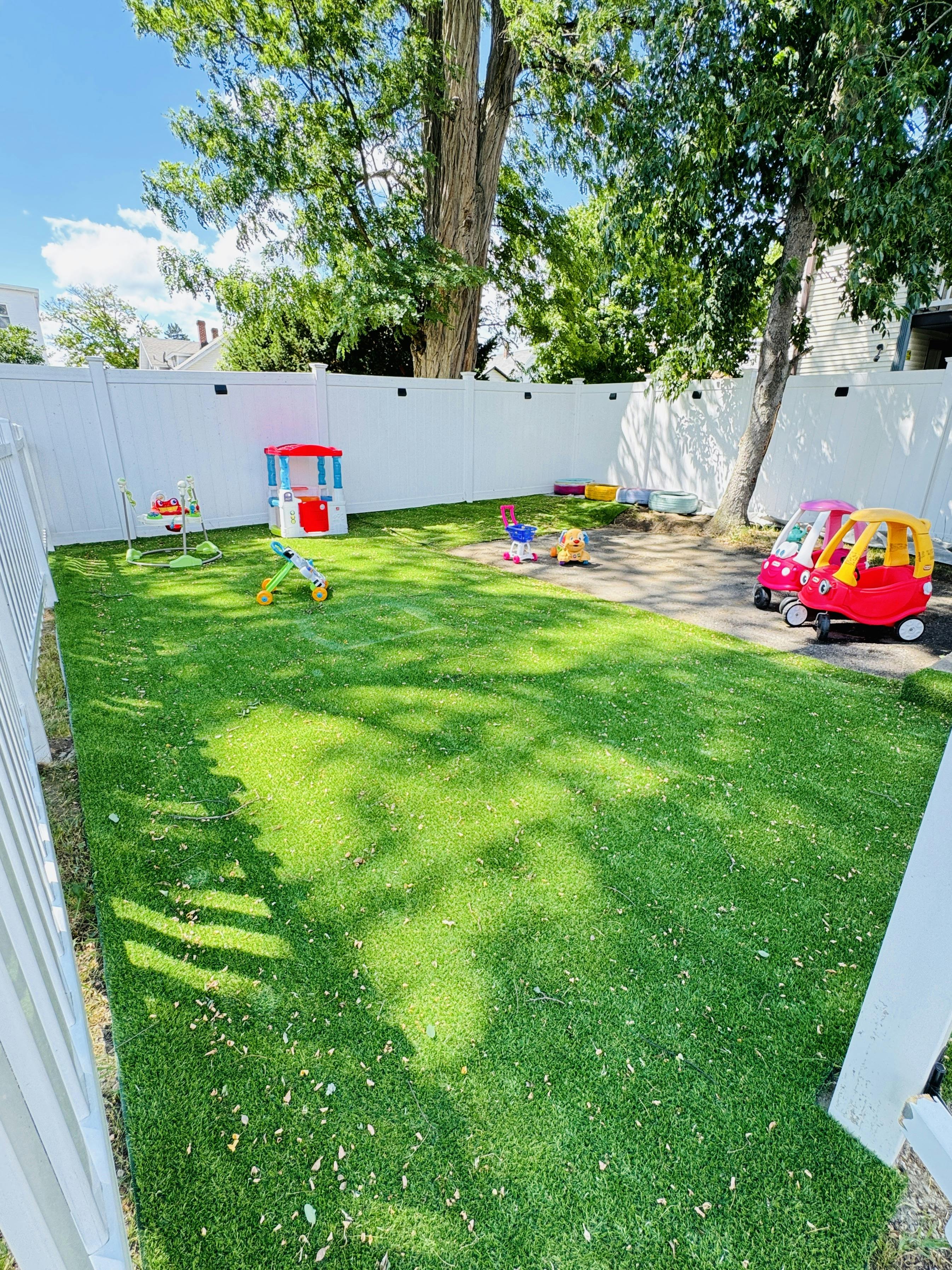 Plastic toys scattered on a grassy backyard play area enclosed by a white fence