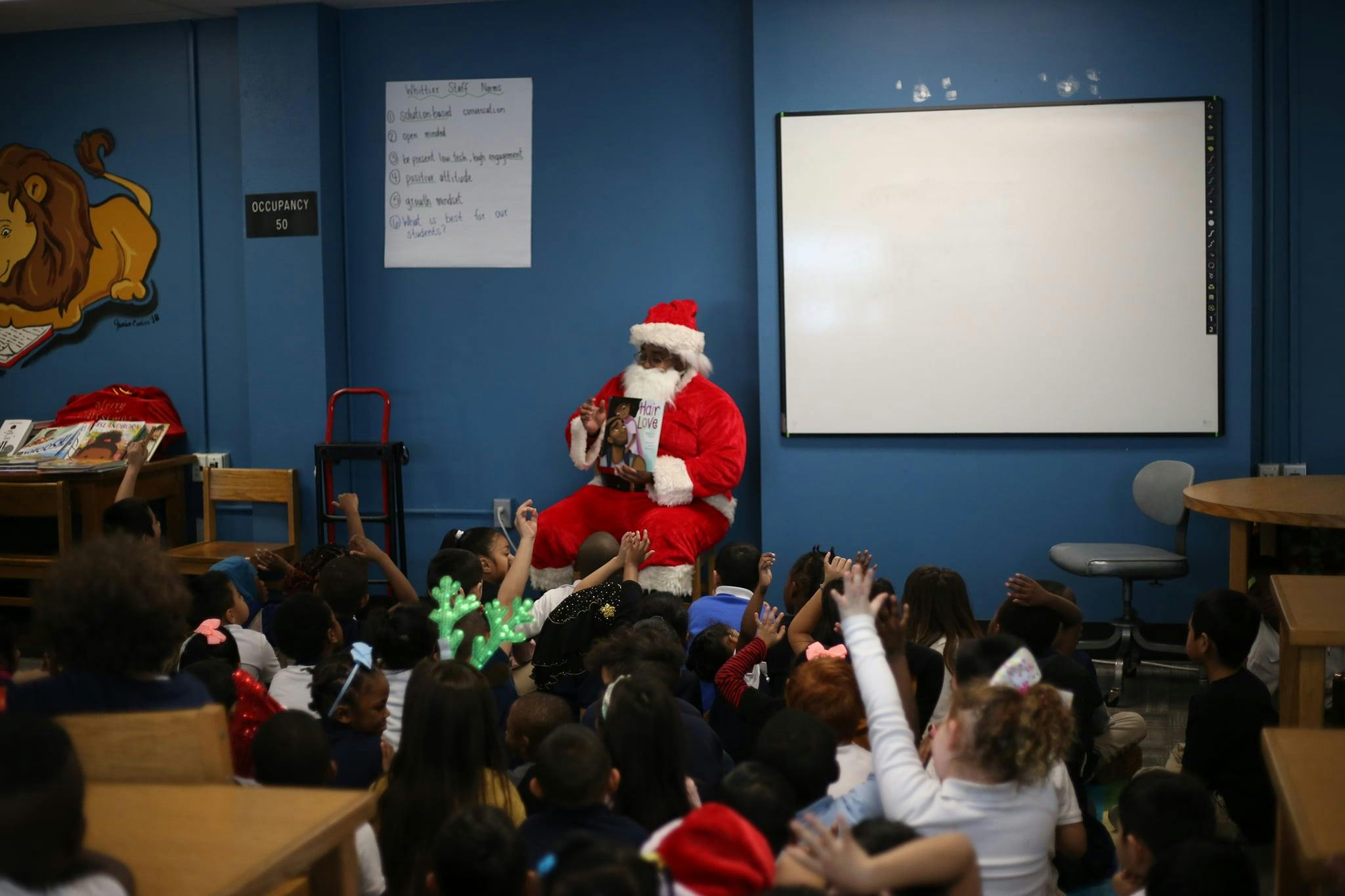 Person in Santa costume sitting with children raising hands in a classroom
